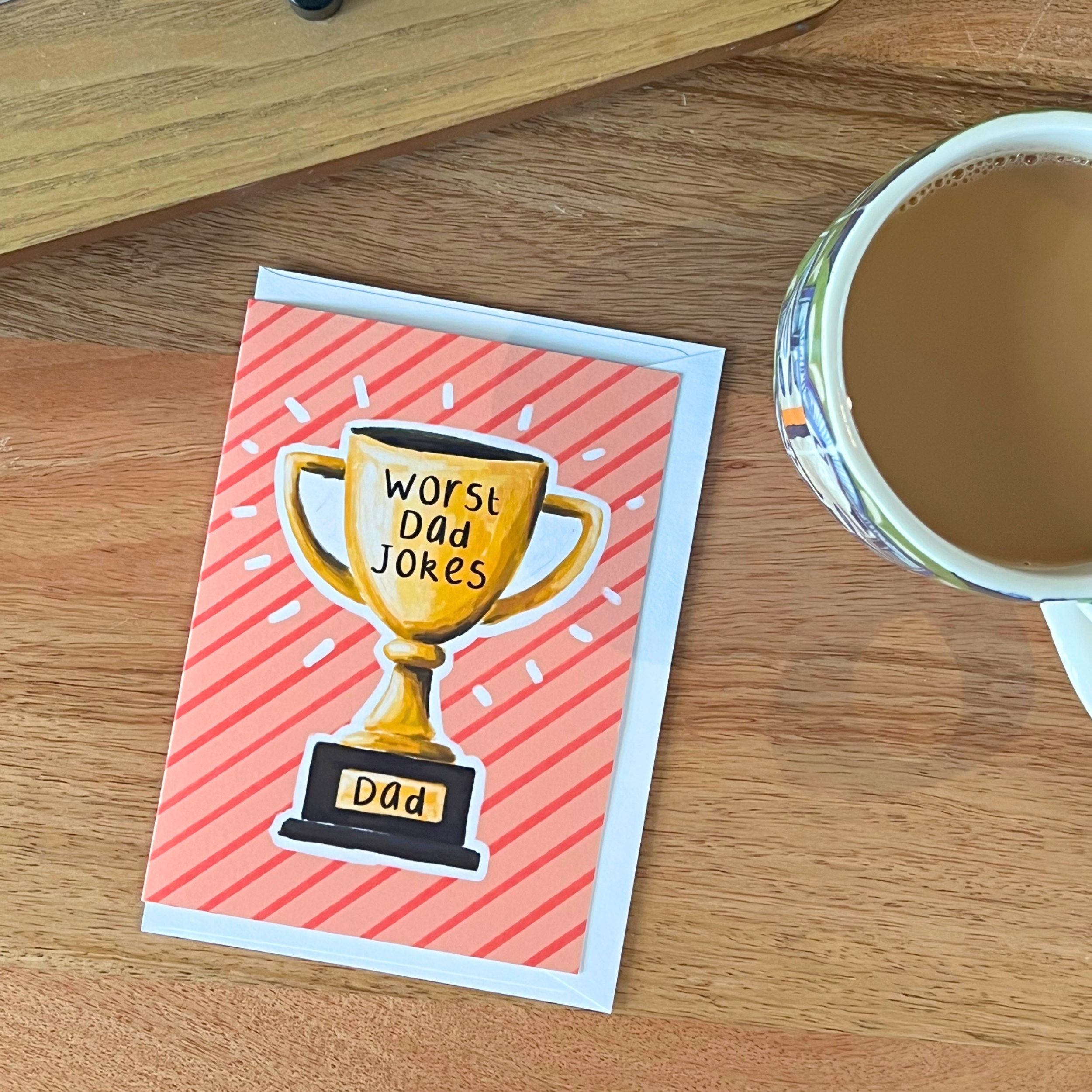 Father’s Day Card with trophy illustration and 'Worst Dad Jokes' text on a wooden surface with a cup of coffee.