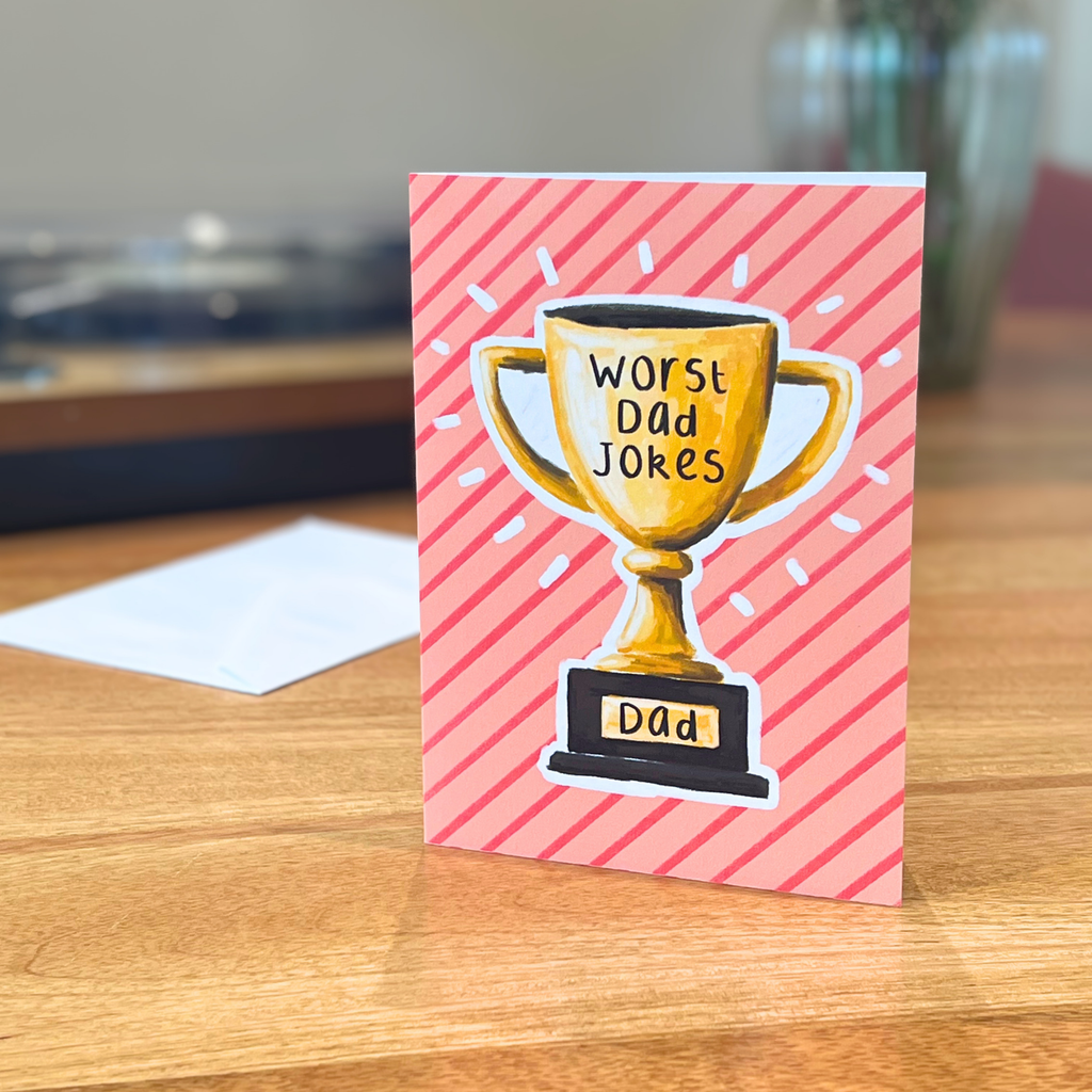 Father’s Day card with a trophy illustration and text 'Worst Dad Jokes' on a pink striped background, placed on a wooden surface.