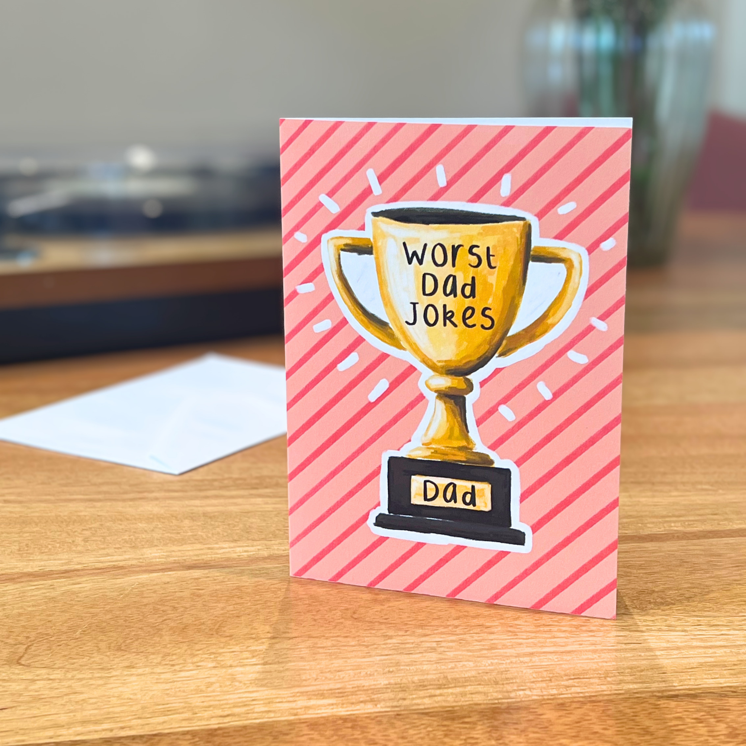 Father’s Day card with a trophy illustration and text 'Worst Dad Jokes' on a pink striped background, placed on a wooden surface.