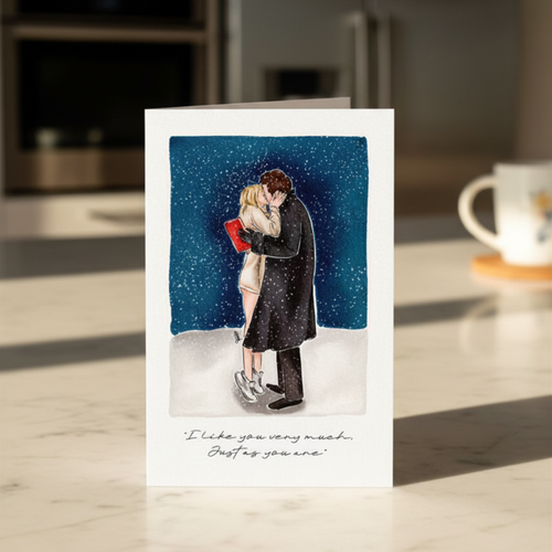 Greeting card with illustration of a couple kissing in the snow with a quote beneath, placed on a kitchen surface.
