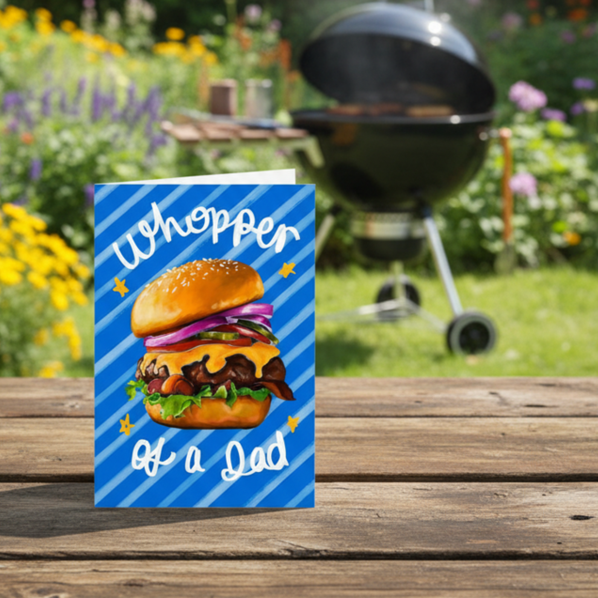 Father’s Day card with Illustration of a burger with text 'Whopper of a Dad' on a blue striped background. Displayed on a a garden table in front of a bbq.