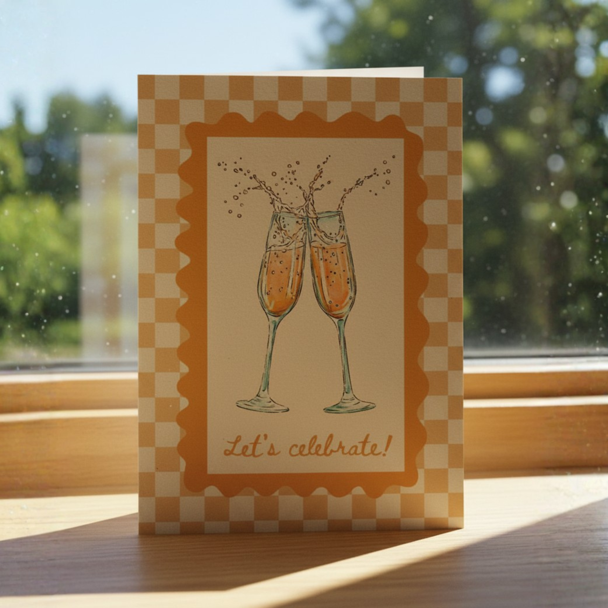 Greeting card with champagne glasses and 'Let's celebrate!' text on a checkered background, placed on a wooden surface with a window in the background.
