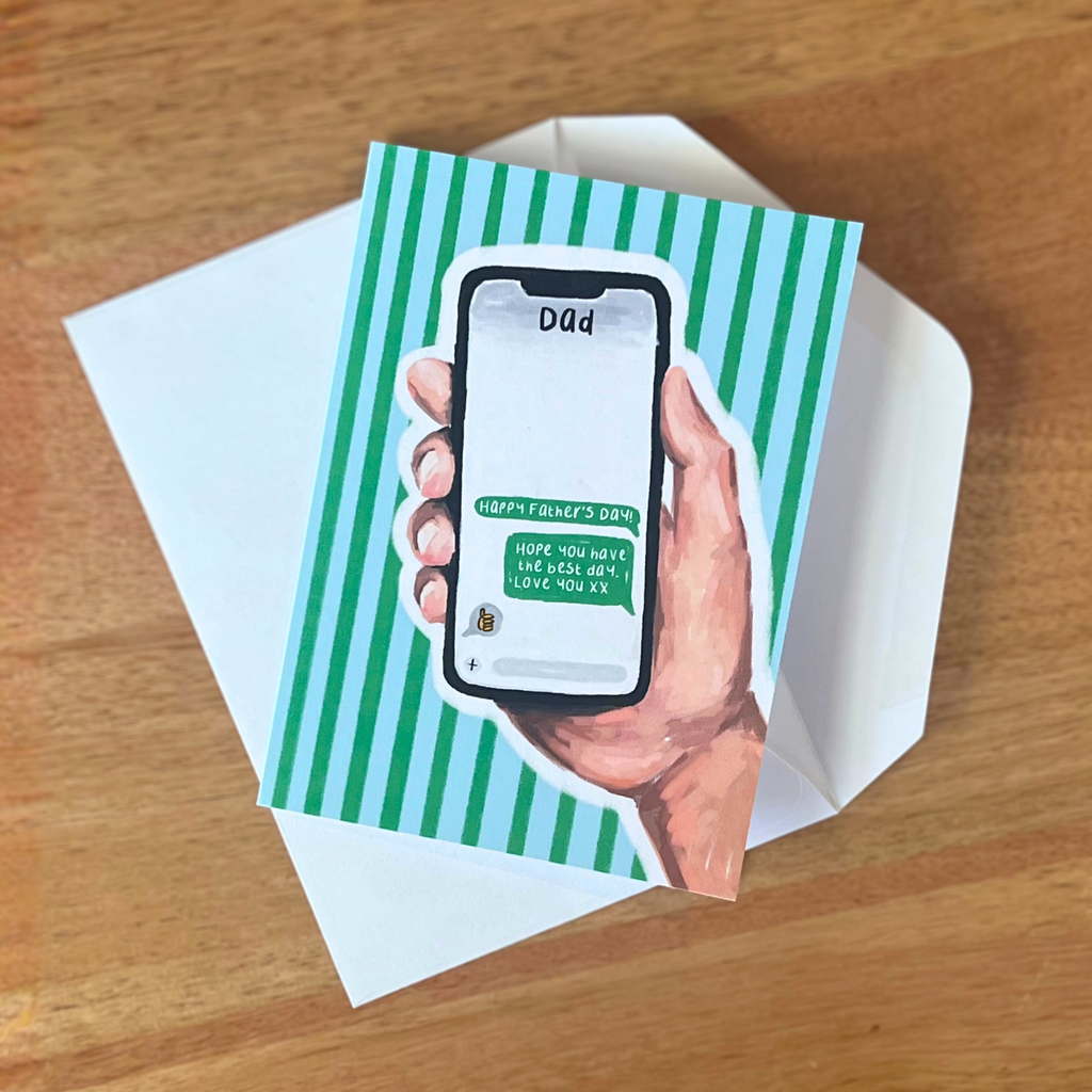 Fathers day card with a hand holding a phone displaying a message on a green striped background, placed on a wooden surface.