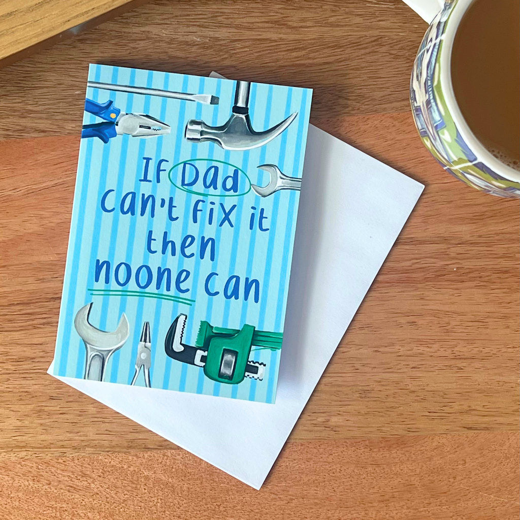 Fathers day card with tools illustration and humorous text on a wooden surface with a cup of coffee.