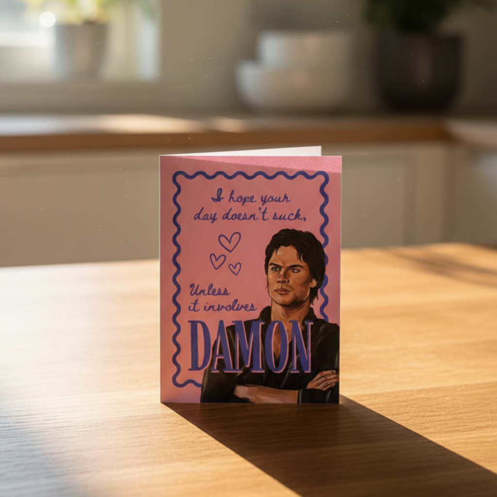 Card with a portrait of a brooding man on a pink background and “i hope your day doesn’t suck unless it involves Damon’ text on a kitchen counter