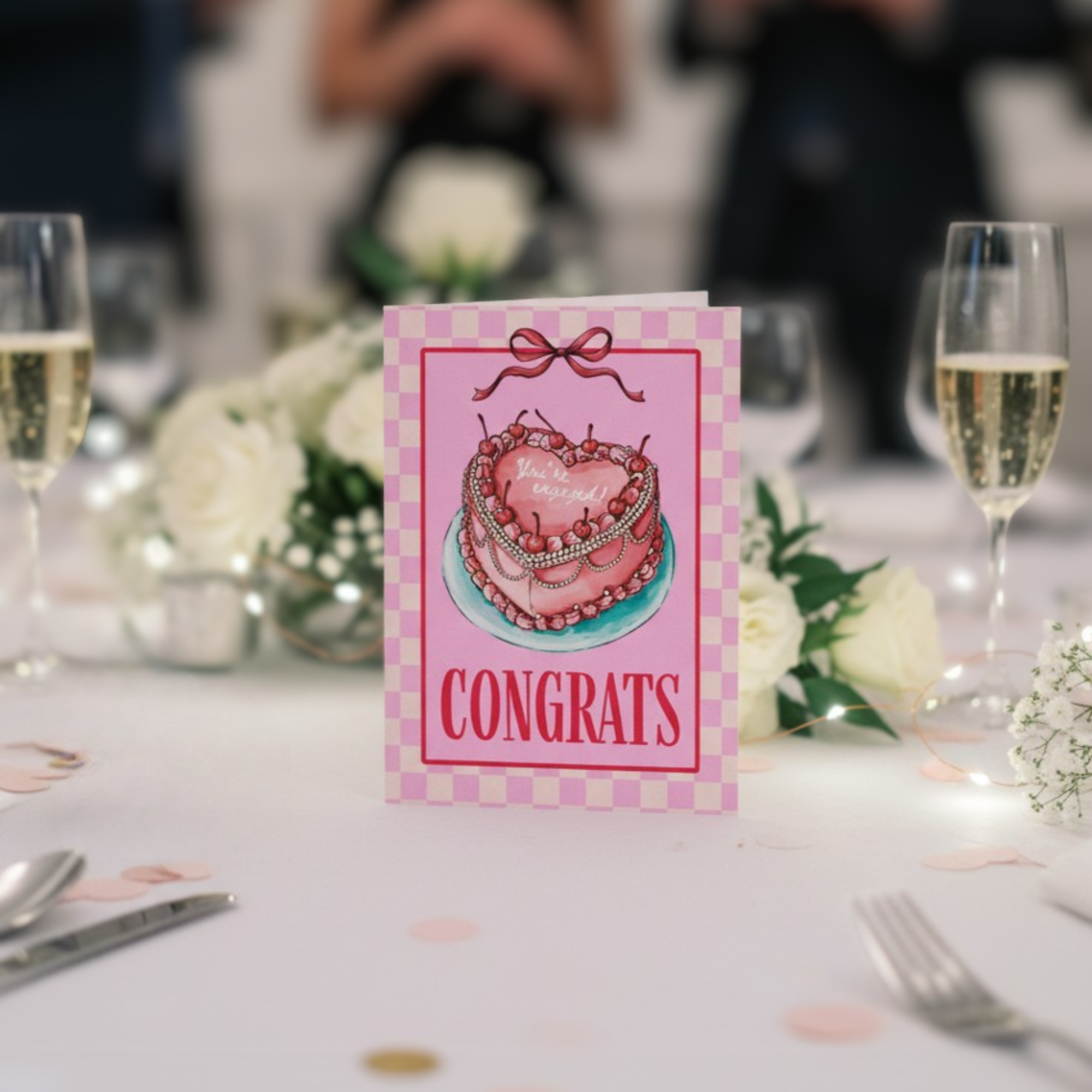 Congrats card with pink heart cake with ‘you’re engaged’ text on a decorated table with flowers and glasses