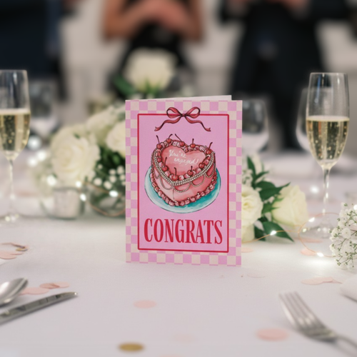 Congrats card with pink heart cake with ‘you’re engaged’ text on a decorated table with flowers and glasses