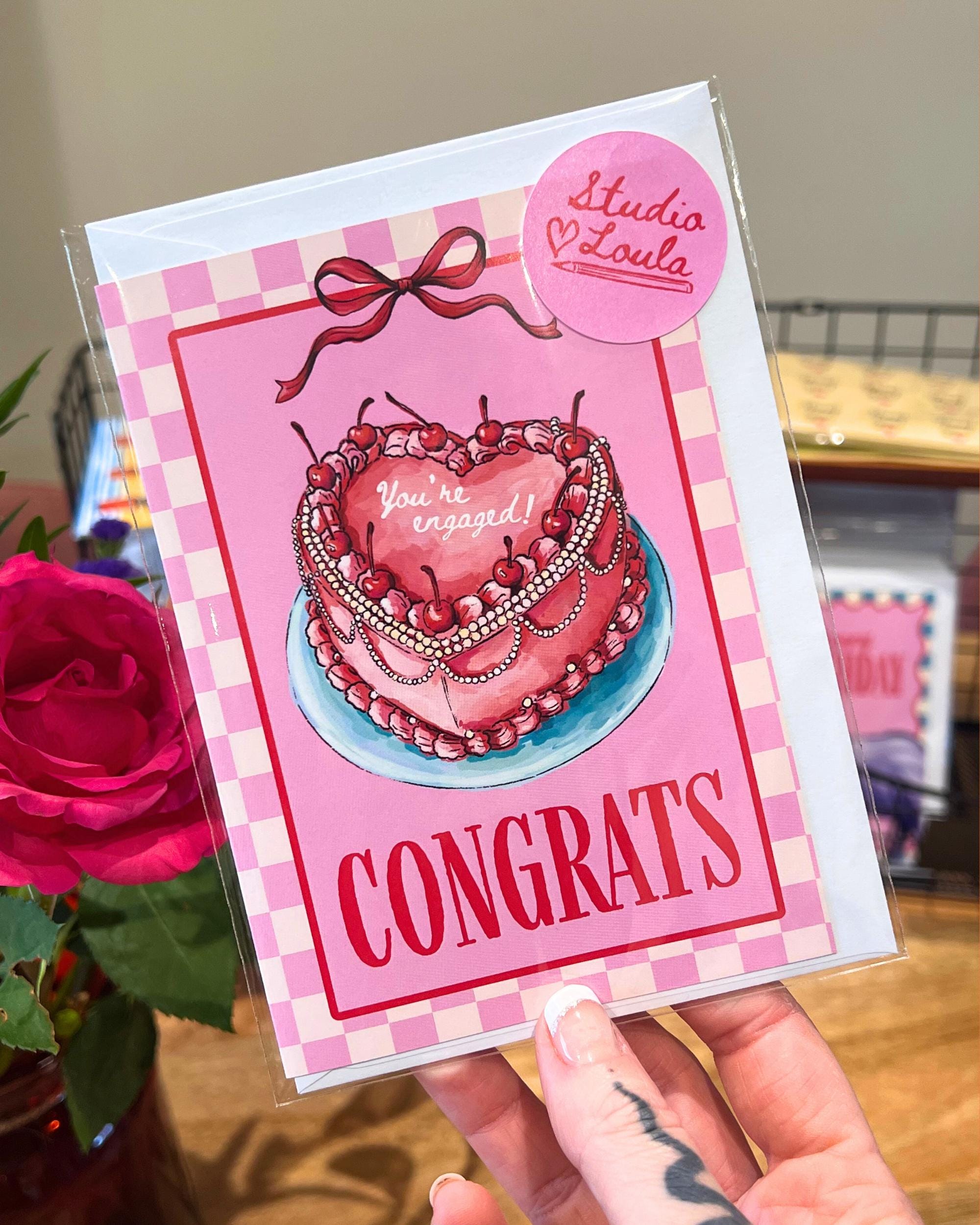 Engagement card with a heart-shaped cake illustration and 'Congrats' text, held by a hand.