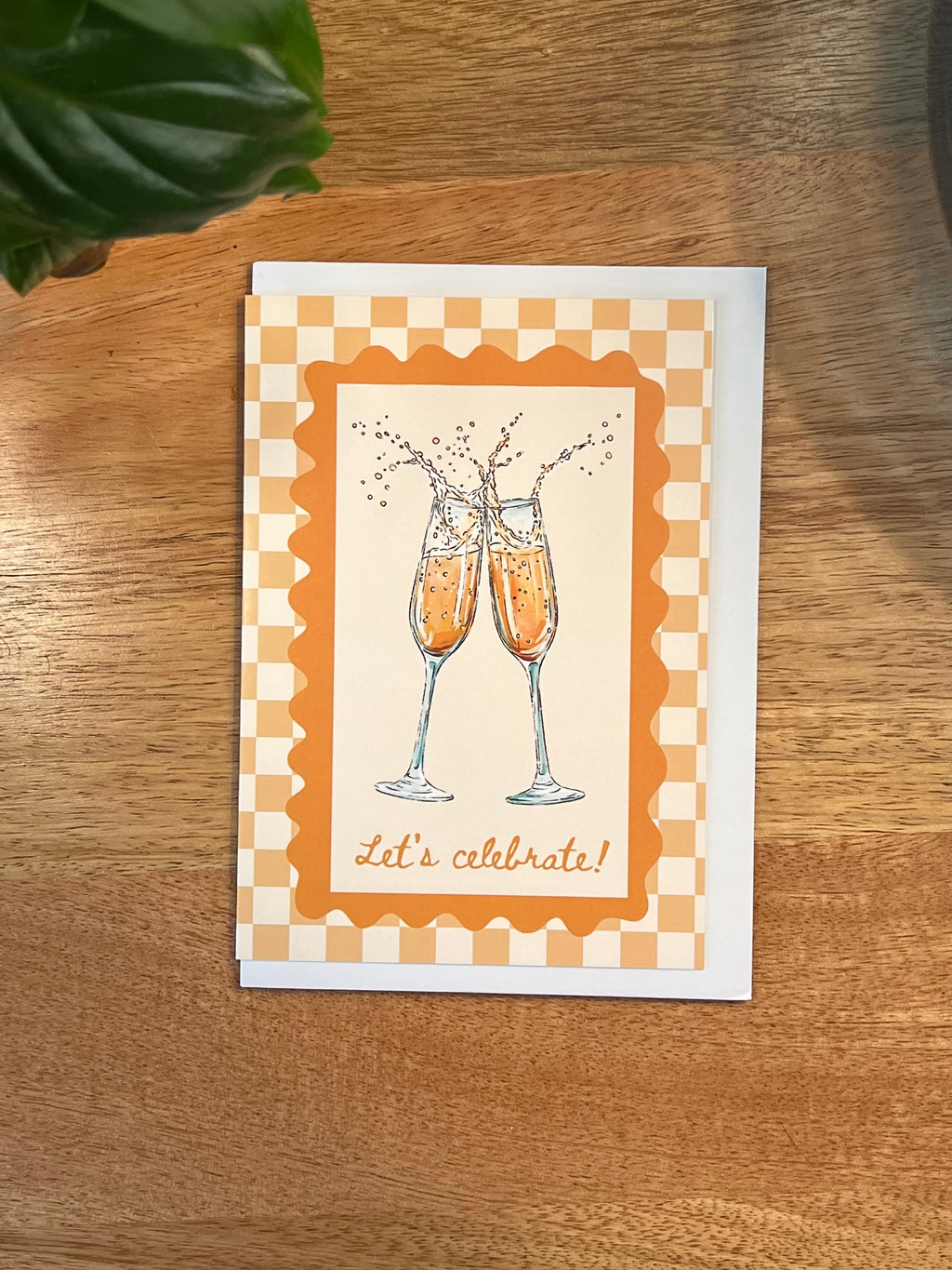 A square greeting card featuring an illustration of two clinking champagne flutes with the words 'let's celebrate!' at the bottom, placed on a wooden tabletop.