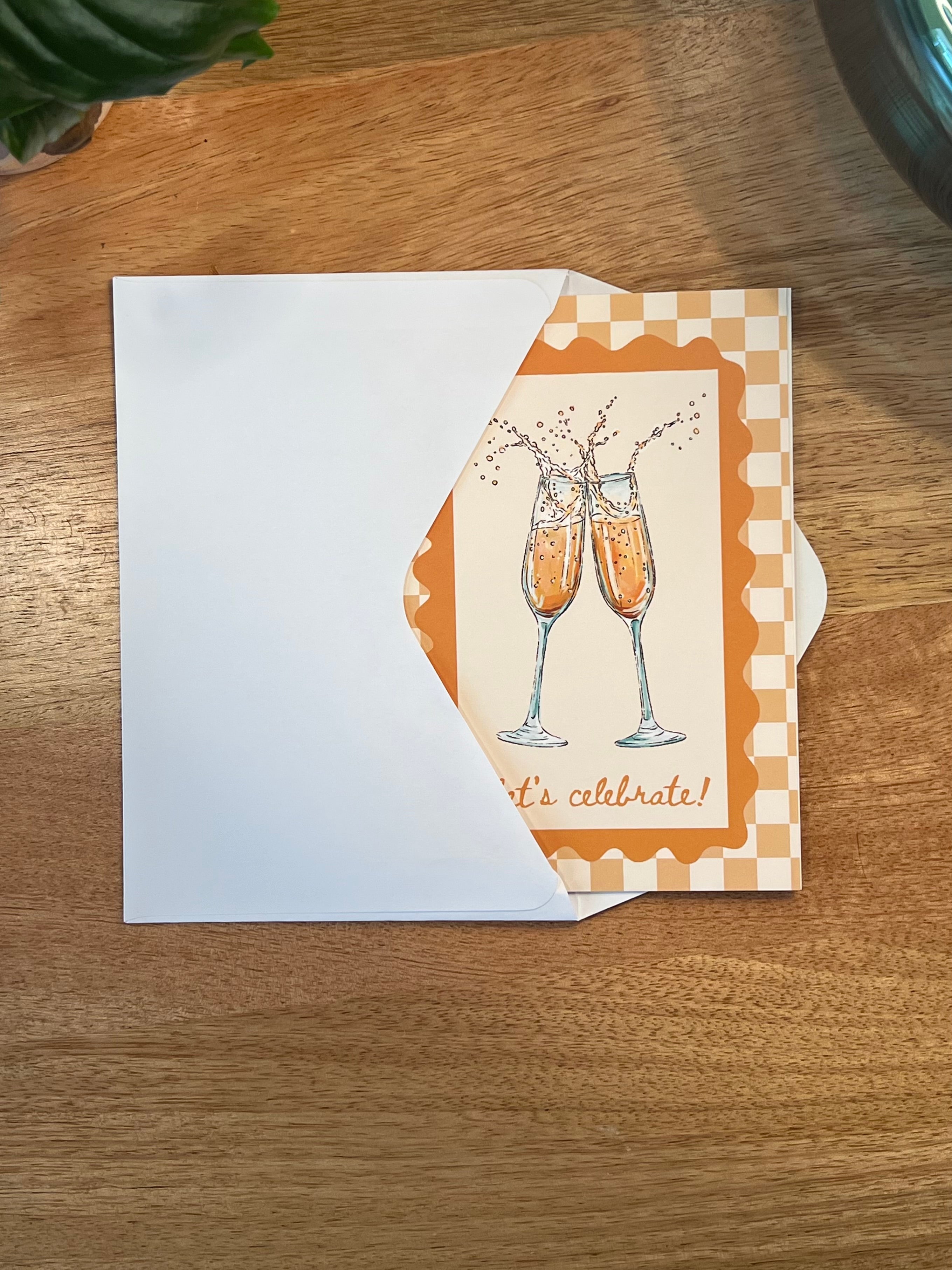 A square greeting card with a white envelope, featuring an illustration of two clinking champagne flutes and the words 'it's celebrate!' on a warm wood tabletop.