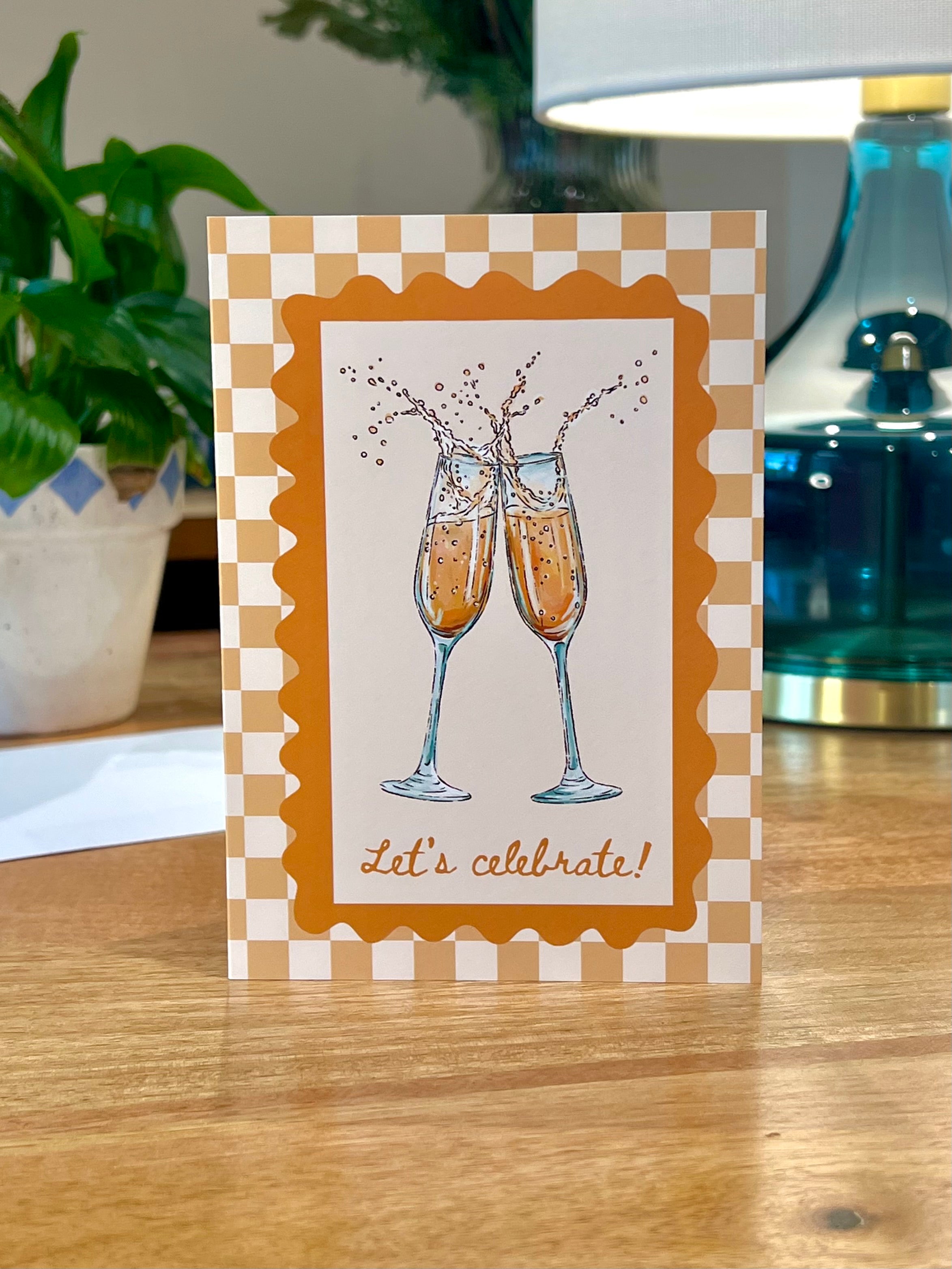 A square greeting card with a checkered border and an illustration of two clinking champagne flutes, standing on a wooden table with a plant and lamp in the background.