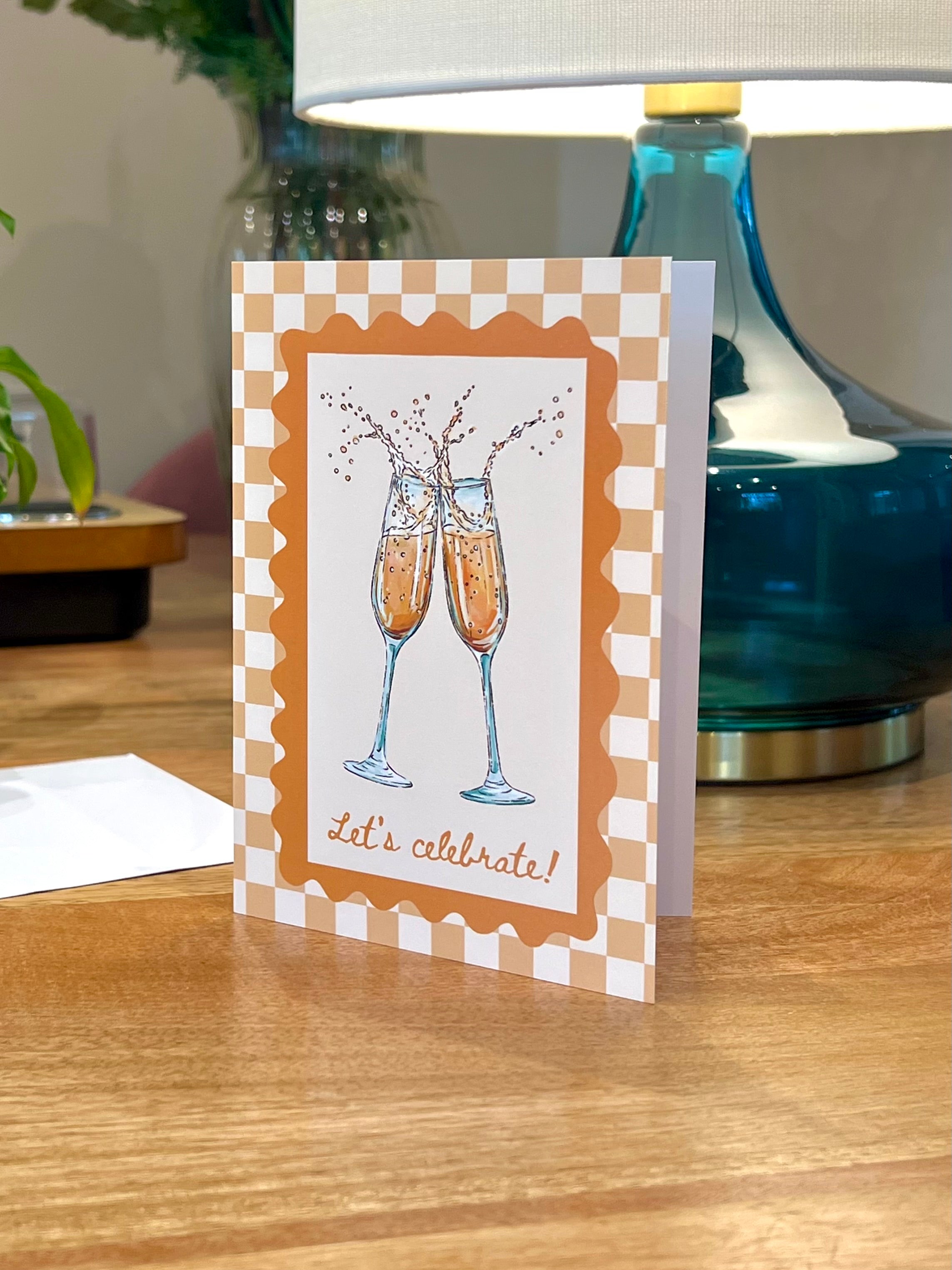 A greeting card standing on a wooden table, featuring an illustration of two champagne flutes with bubbles and the words 'let's celebrate!' in brown script. The card has a white and tan checkered border and is photographed in a real indoor setting with a lamp and plant in the background.