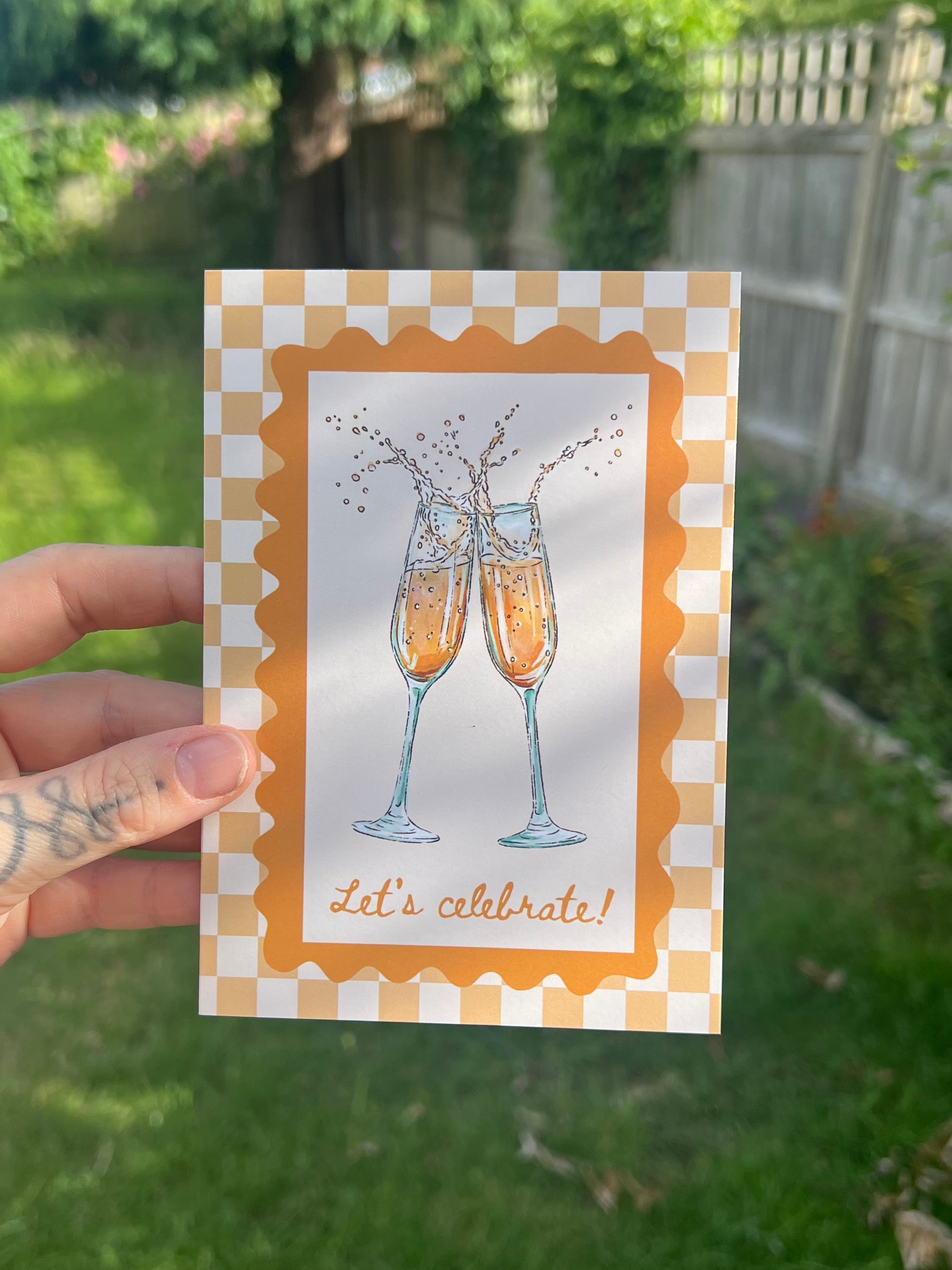 A hand holds a square greeting card outdoors, with a blurred garden background. The card features an illustration of two clinking champagne glasses with the text 'Let's celebrate!' and a yellow-and-white checkered border.