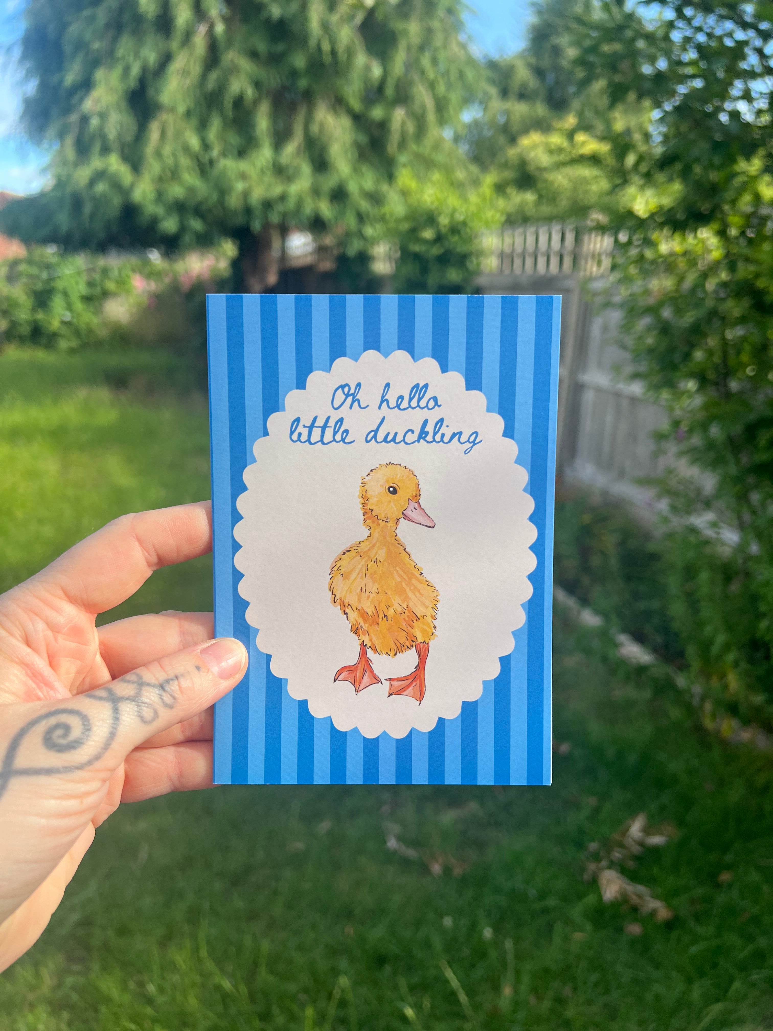 A hand with a visible tattoo holds a square greeting card outdoors. The card has a blue and white striped border, a white scalloped center, and a yellow duck illustration with the text 'Oh, little little duckling'.