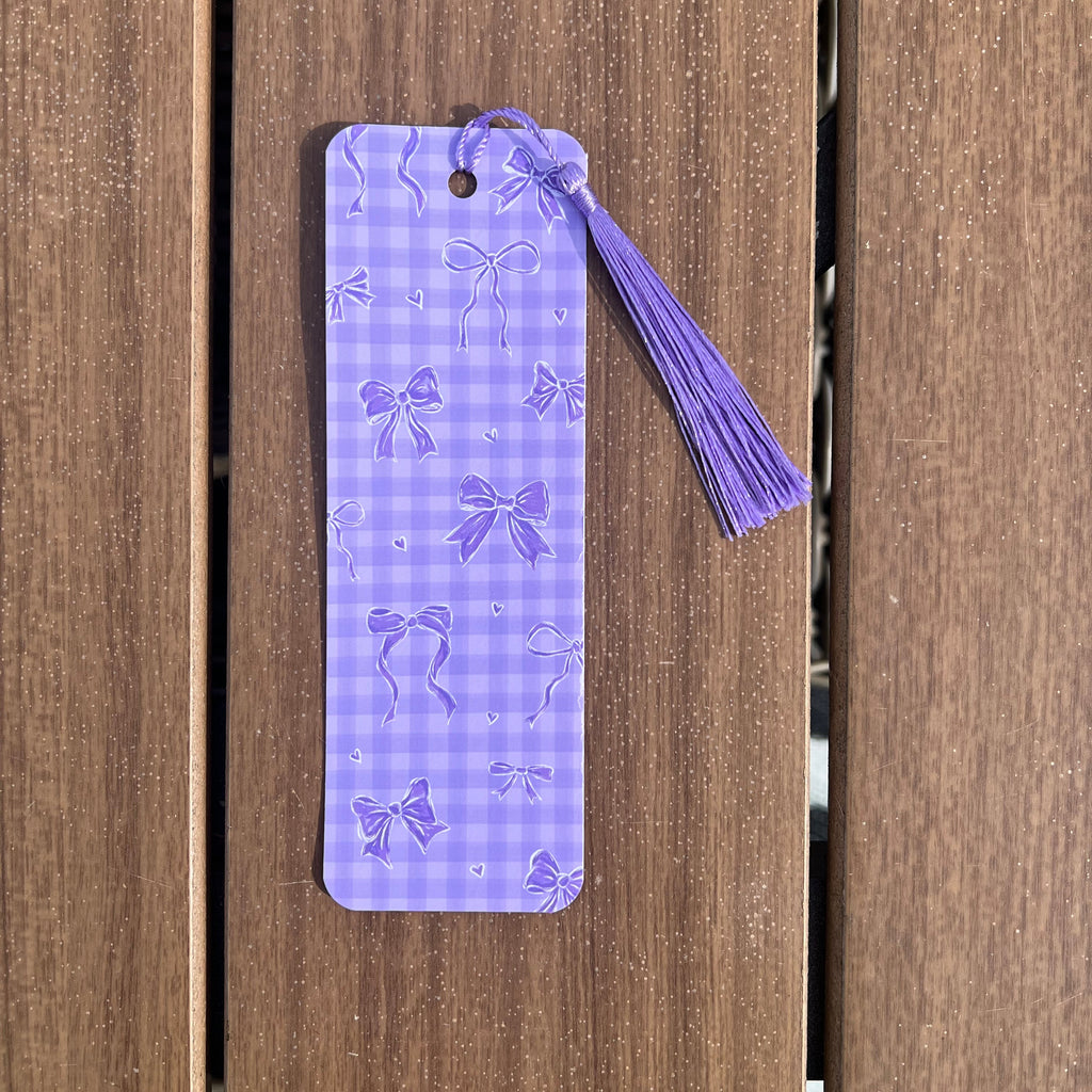 A lavender purple gingham bookmark with embossed bow patterns and a matching tassel, shown against a brown wooden plank background.