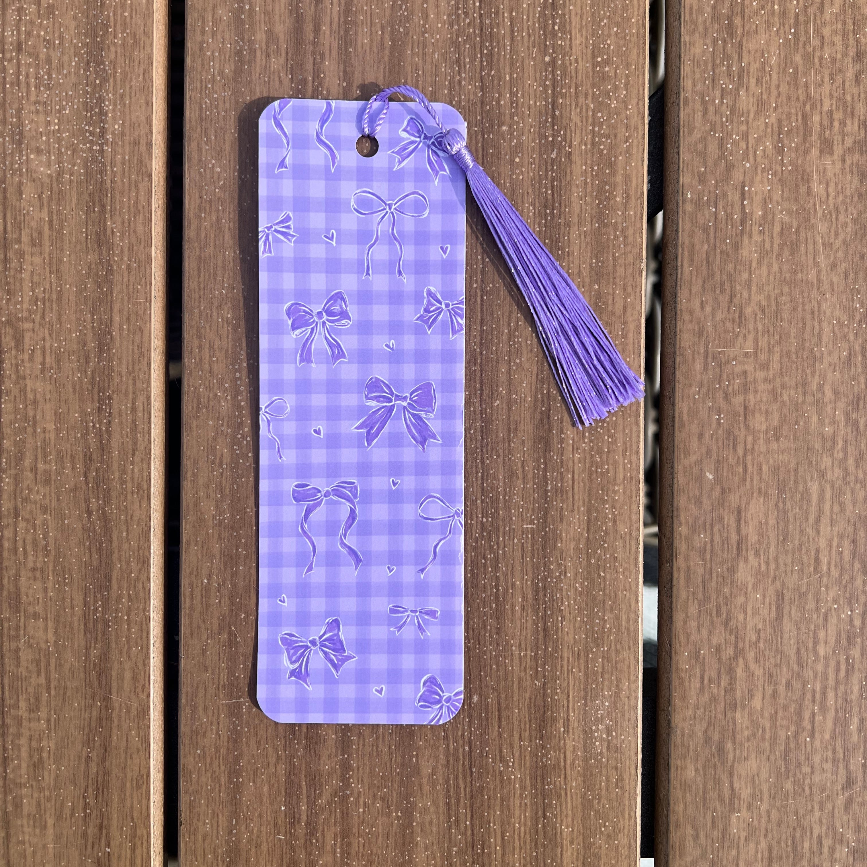 A lavender purple gingham bookmark with embossed bow patterns and a matching tassel, shown against a brown wooden plank background.