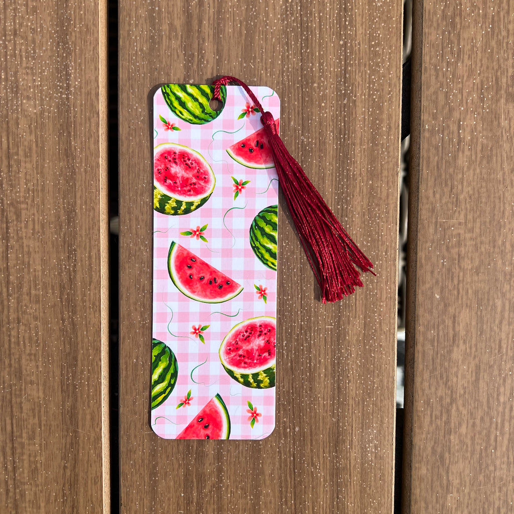 A rectangular paper bookmark with a watermelon slice pattern on a pink gingham background, featuring a dark red tassel at the top, shown against a brown wooden plank background.