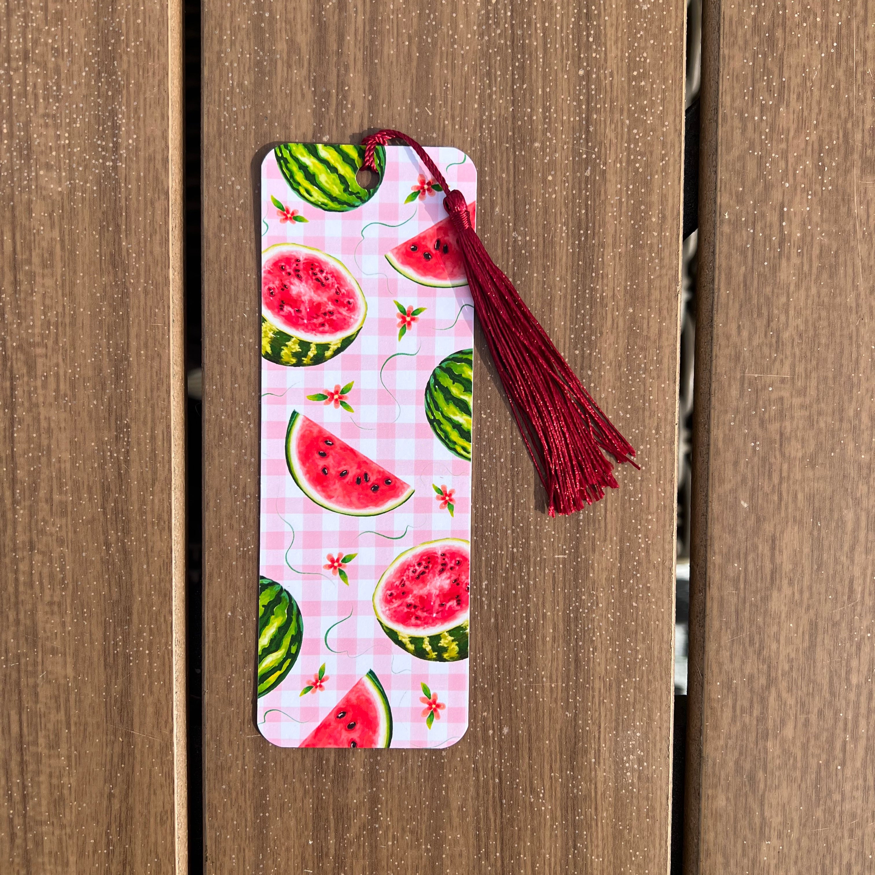 A rectangular paper bookmark with a watermelon slice pattern on a pink gingham background, featuring a dark red tassel at the top, shown against a brown wooden plank background.