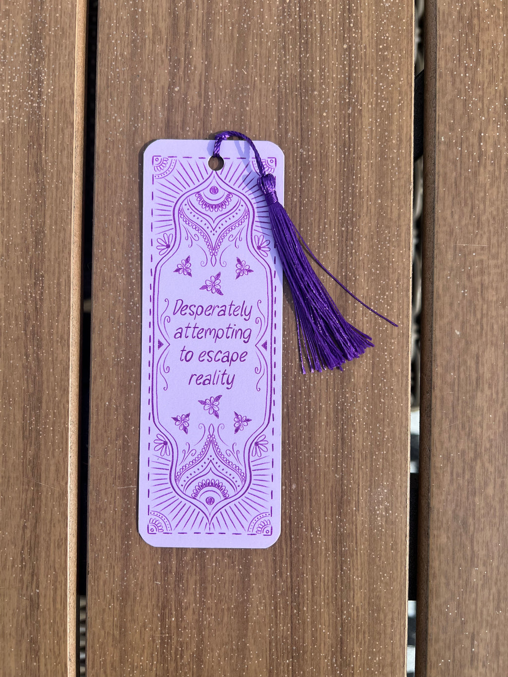 A lavender paper bookmark with purple mandala-style illustrations and the phrase 'Desperately attempting to escape reality' in script, attached to a purple tassel, photographed against a brown wooden slat background.