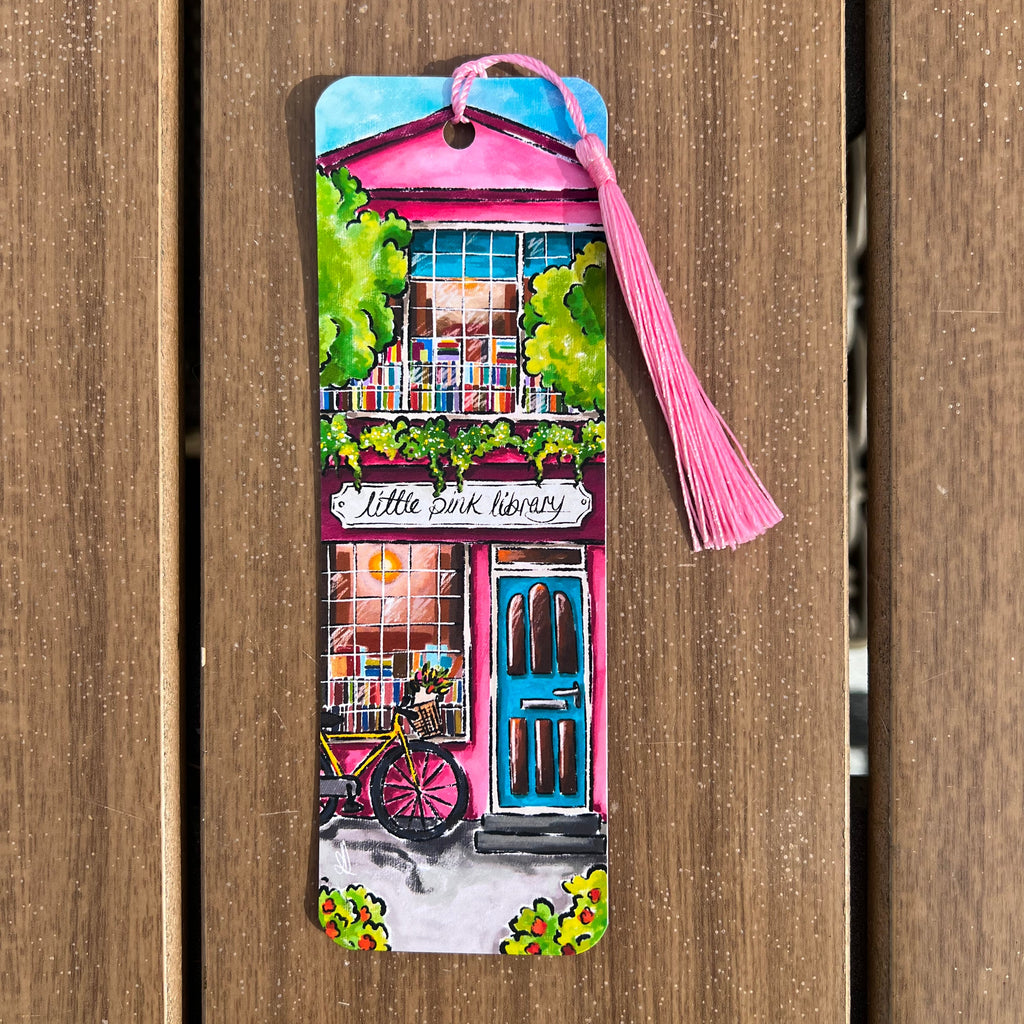 A colorful hand-painted wooden bookmark shaped like a bookshop storefront with a pink tassel, photographed against a brown wooden plank background.