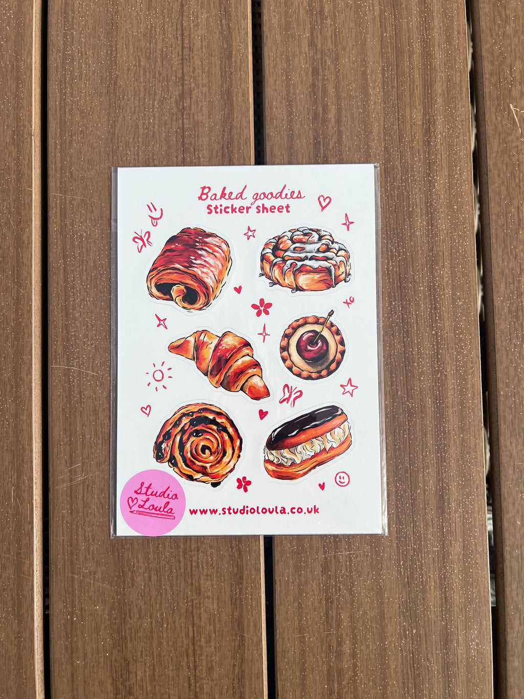 A sticker sheet featuring illustrated baked goods, including a croissant, cinnamon roll, pastry, and donut, placed on a brown wooden surface. The card has red text reading 'Bakery treats' and a pink circular badge with website text.