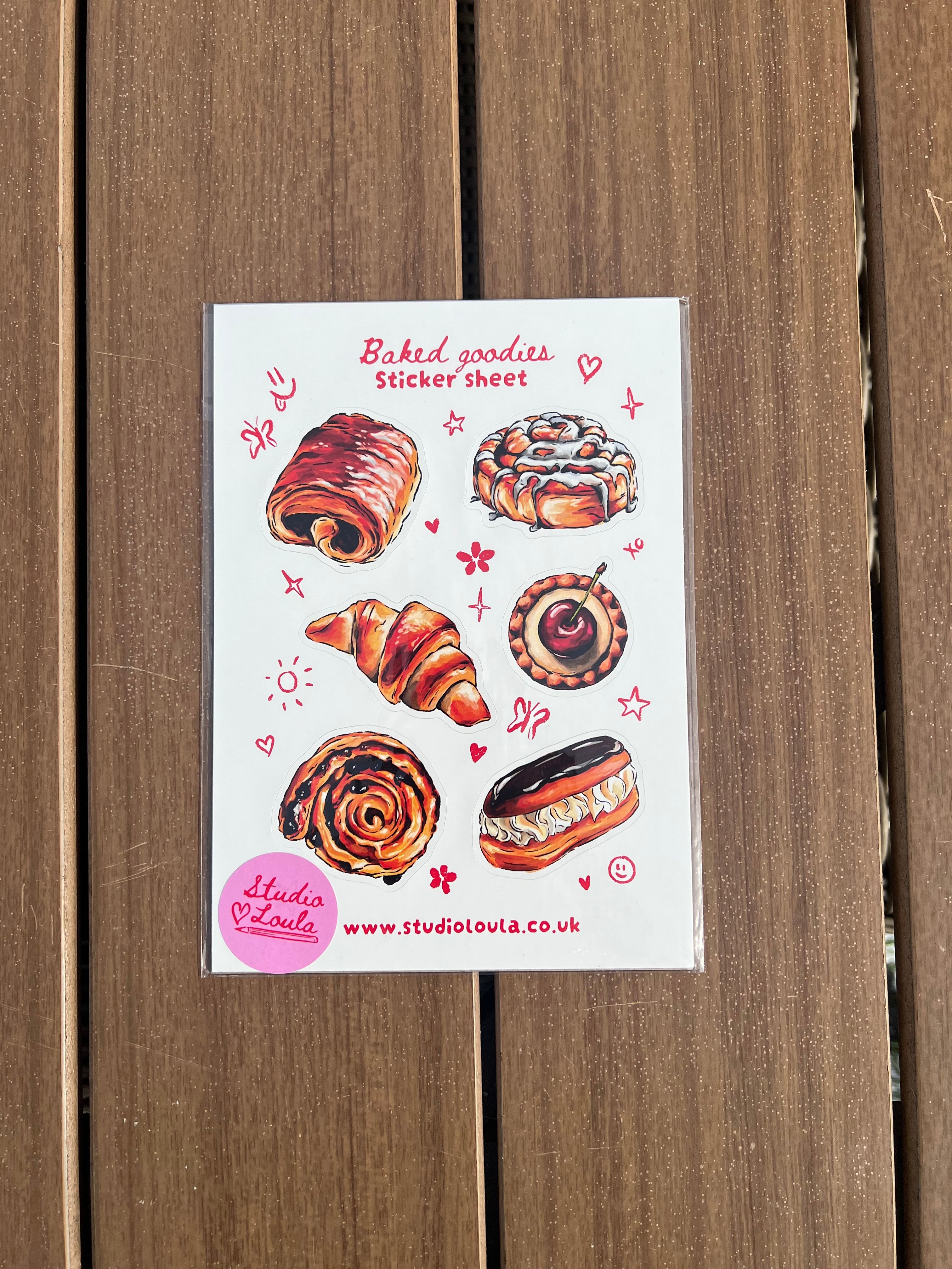 A sticker sheet featuring illustrated baked goods, including a croissant, cinnamon roll, pastry, and donut, placed on a brown wooden surface. The card has red text reading 'Bakery treats' and a pink circular badge with website text.