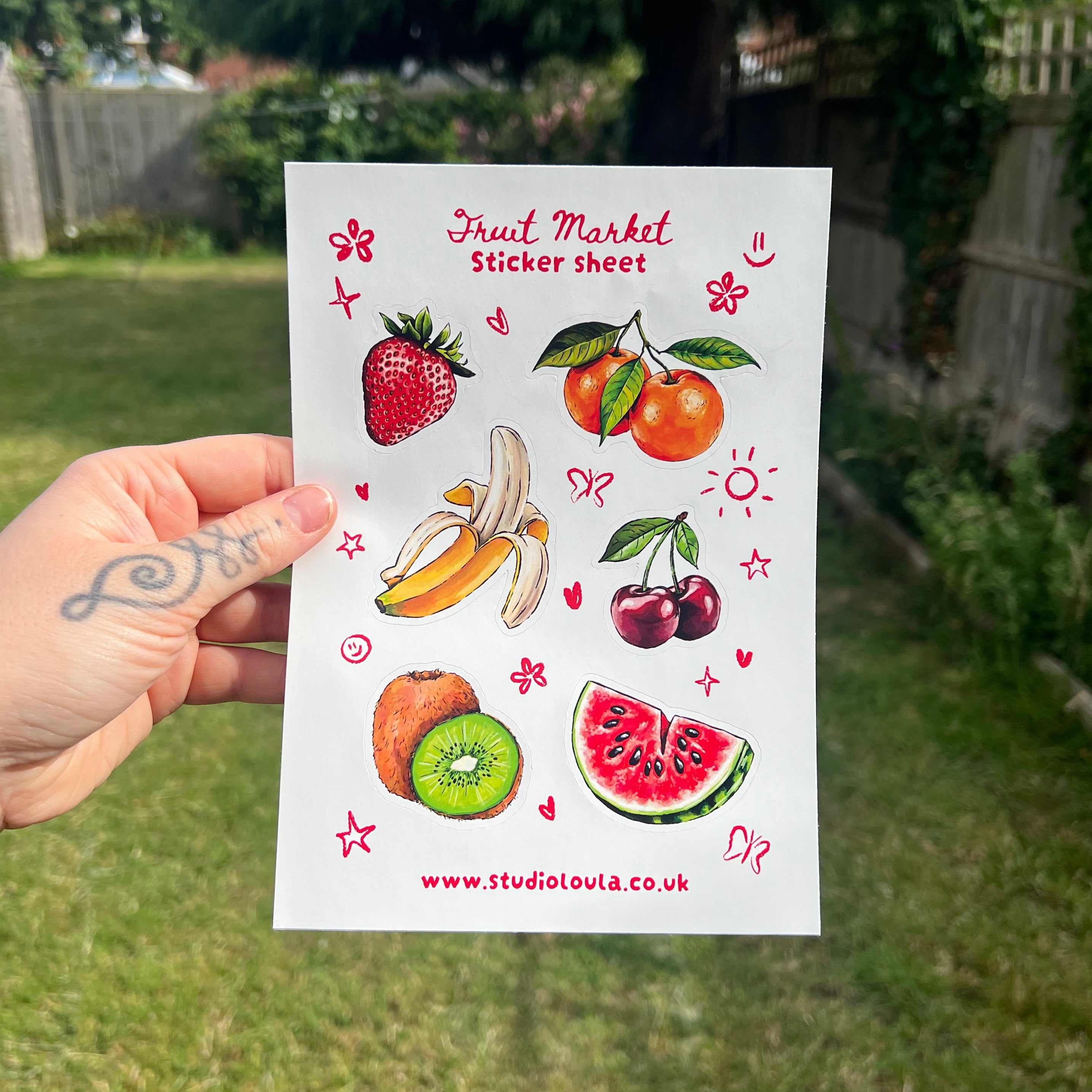 A hand holds a white sticker sheet featuring colorful watercolor illustrations of various fruits, including strawberries, oranges, bananas, cherries, kiwi, and watermelon, against a blurred green garden background.