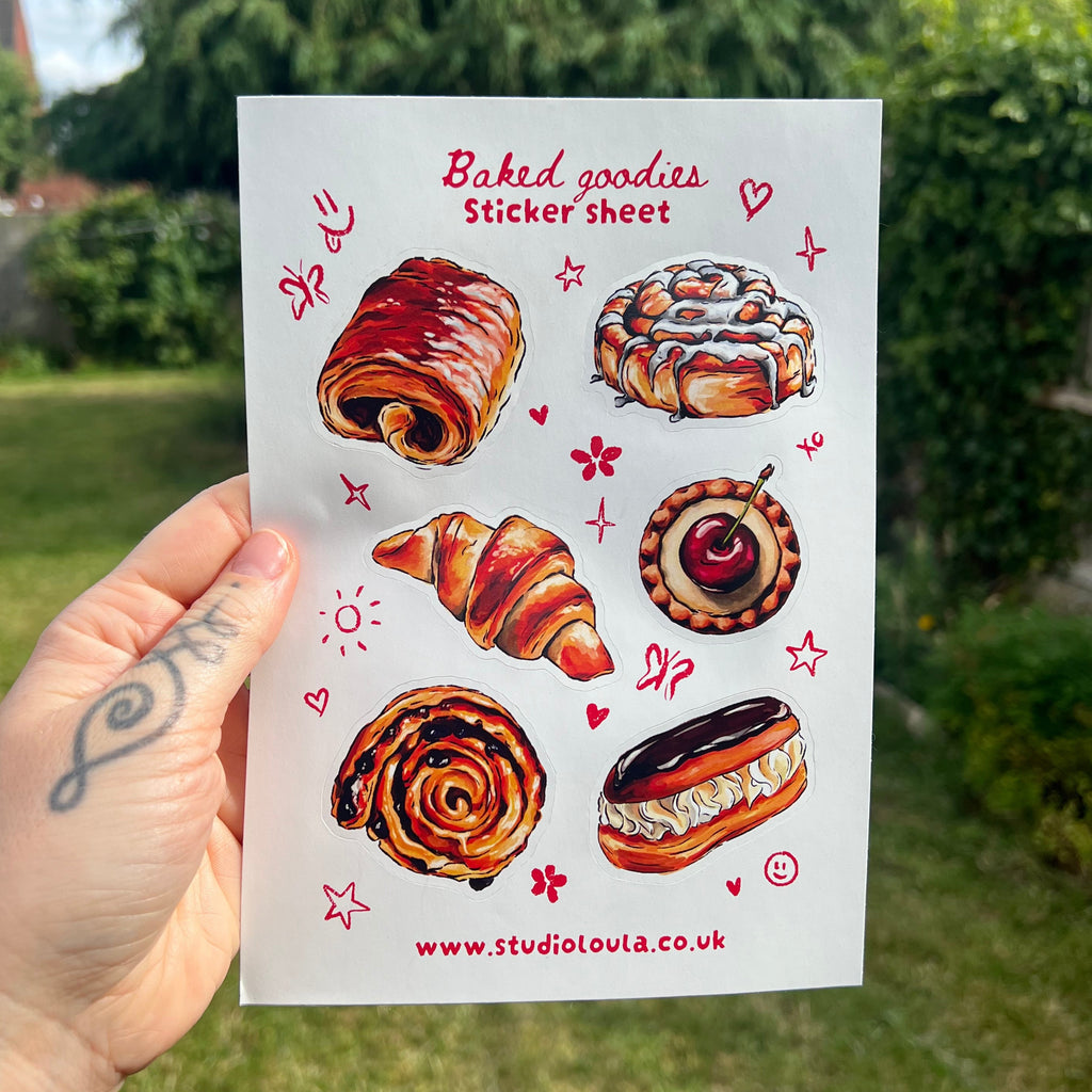 A hand holds a white sticker sheet featuring illustrated baked goods like pastries, croissants, and cakes, with red text reading 'Baked goodies Sticker sheet' and the website 'www.studiovlou.