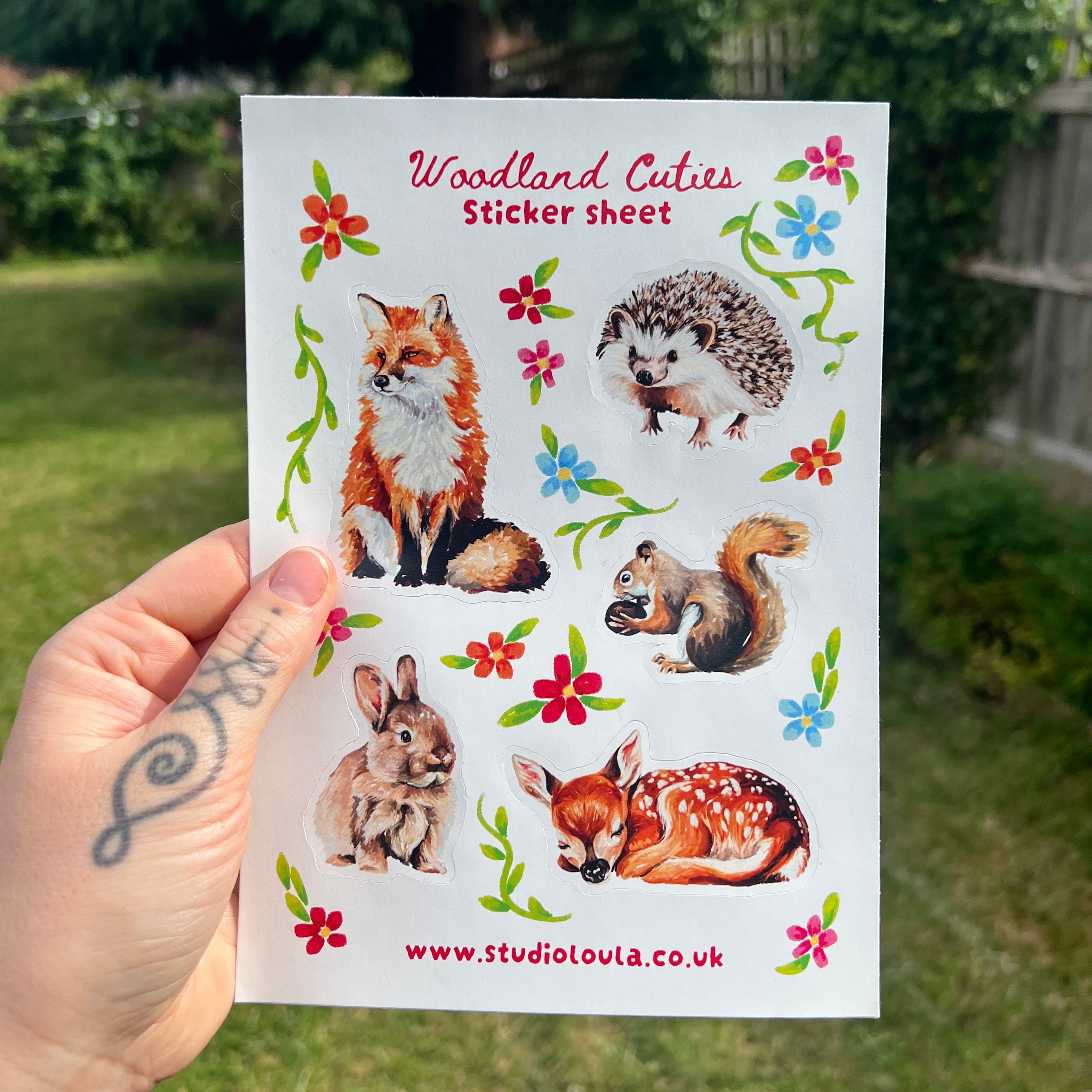 A hand holds a white sticker sheet featuring woodland animal illustrations, including a fox, hedgehog, squirrel, rabbit, and deer, with red and blue flowers and green leaves. The sheet is photographed outdoors against a blurred green garden background.