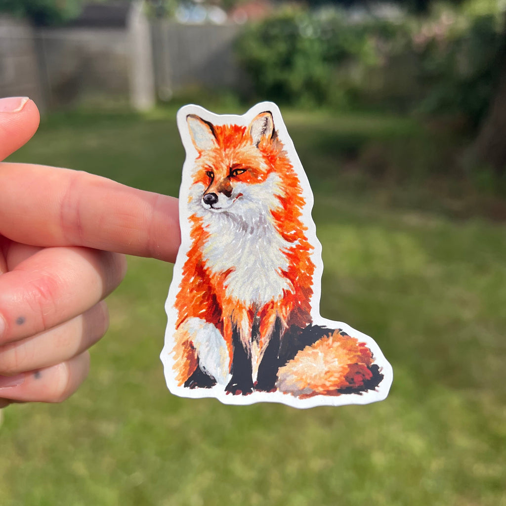 A hand holds a die-cut sticker of a red fox with orange, white, and black fur against a blurred green grassy background.