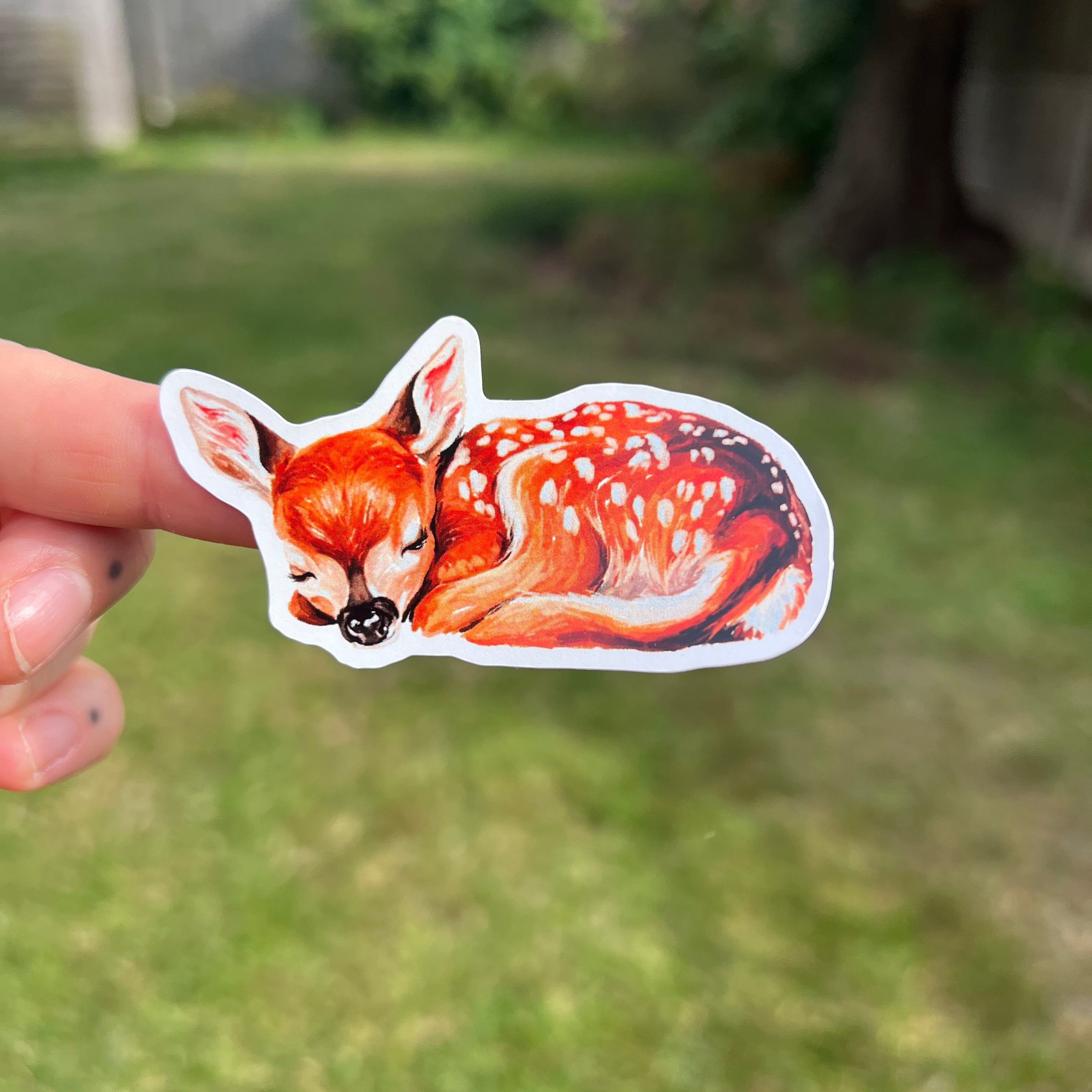 A hand holds a die-cut sticker of a sleeping fawn with orange and white fur spots, set against a blurred green grassy backyard background.