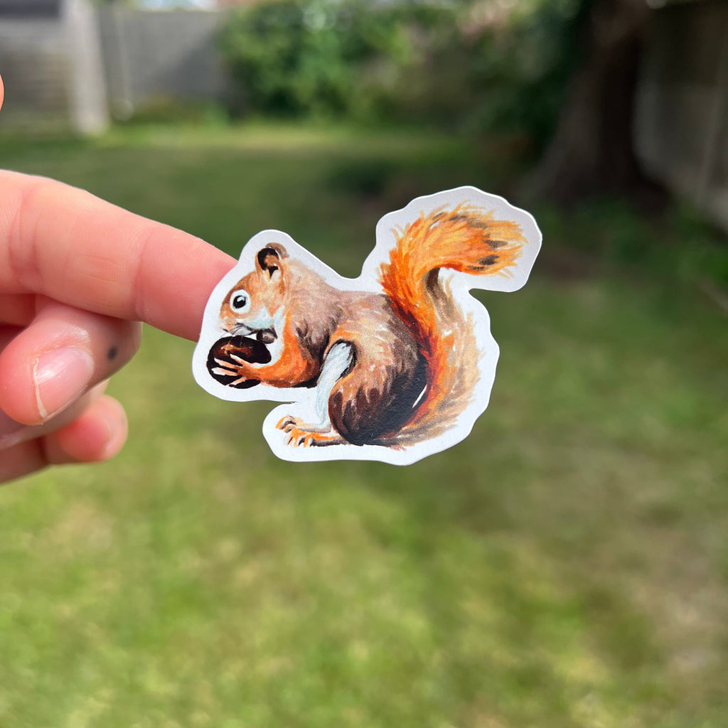 A hand holds a die-cut sticker of a brown and orange squirrel holding a nut, with a blurred green grassy backyard in the background.