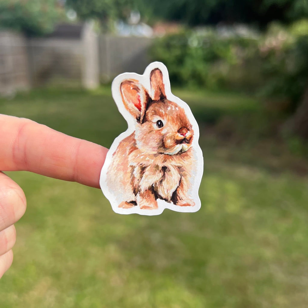 A die-cut sticker of a brown rabbit held by a person's fingers against a blurred green backyard background.