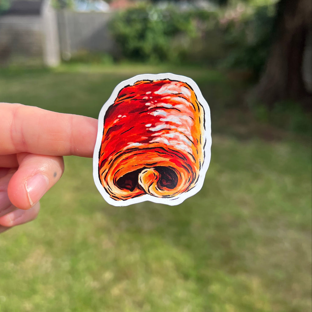 A hand holding a die-cut sticker of a swirl pastry, with a blurred green grassy backyard in the background.