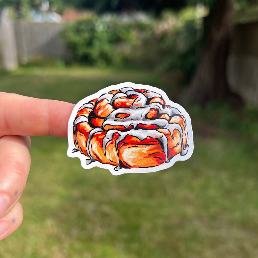A hand holds a die-cut sticker featuring a detailed illustration of a glazed cinnamon roll, set against a blurred outdoor backyard background.