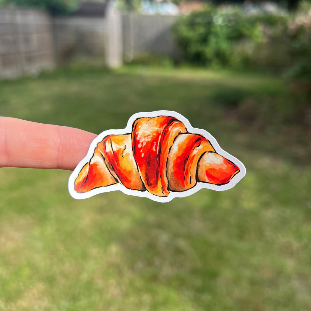 A die-cut vinyl sticker of a watercolor croissant held by a person's finger against a blurred green grassy backyard background.