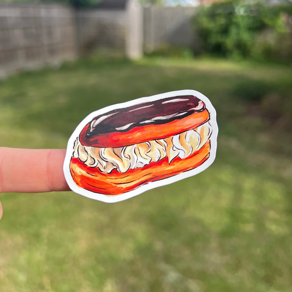 A die-cut sticker of a chocolate-covered pastry filled with cream, held by a person's finger against a blurred green grassy backyard background.