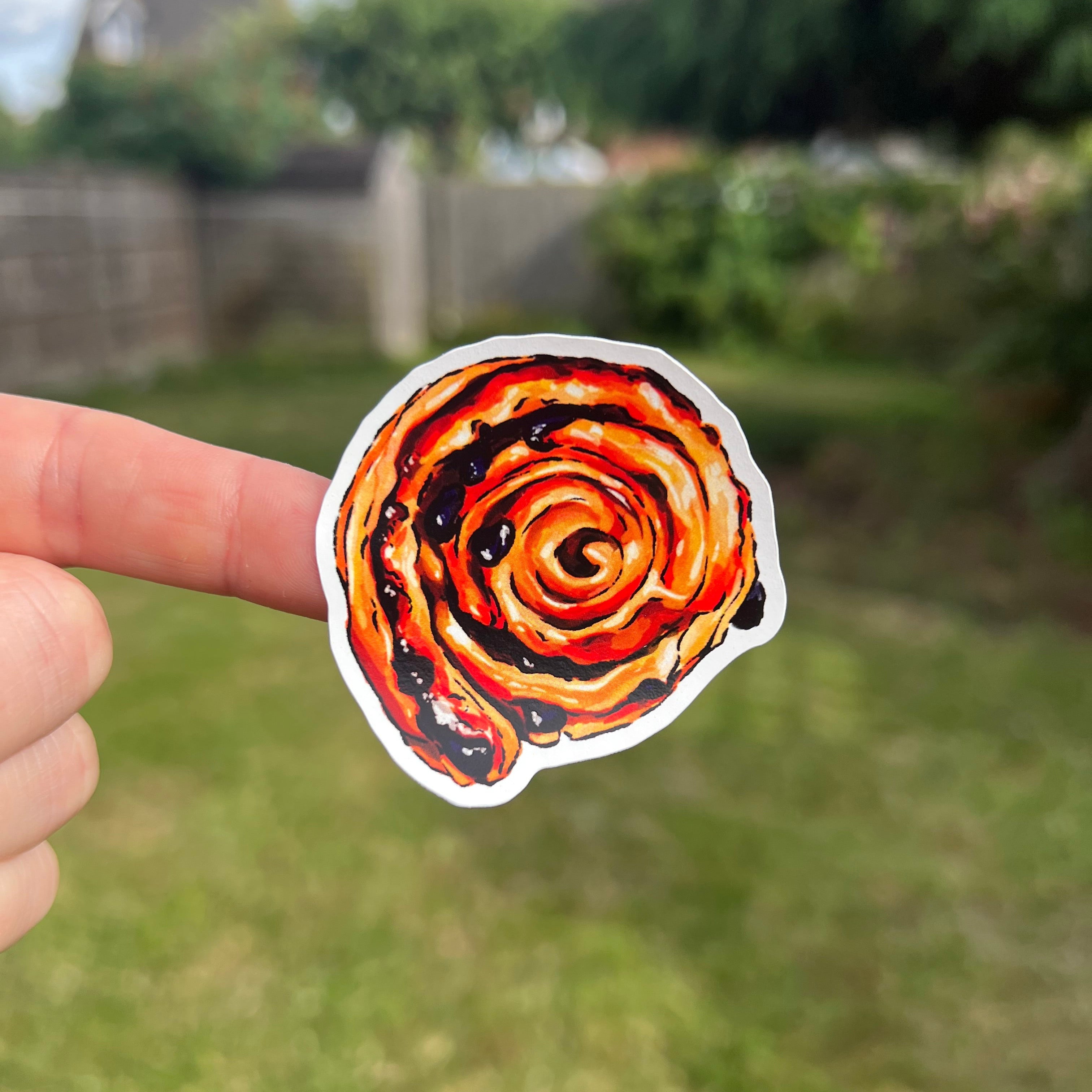 A hand holding a die-cut sticker featuring a pain aux raisin pastry, with a blurred green backyard in the background.