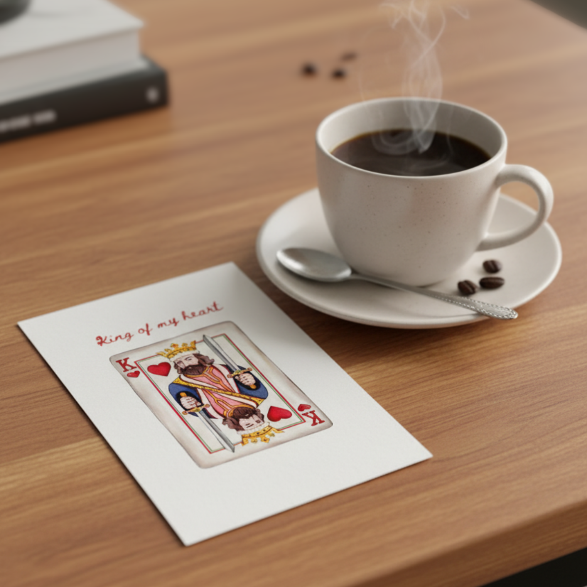 White mug with steaming coffee on a wooden table next to a greeting card featuring king of hearts design 