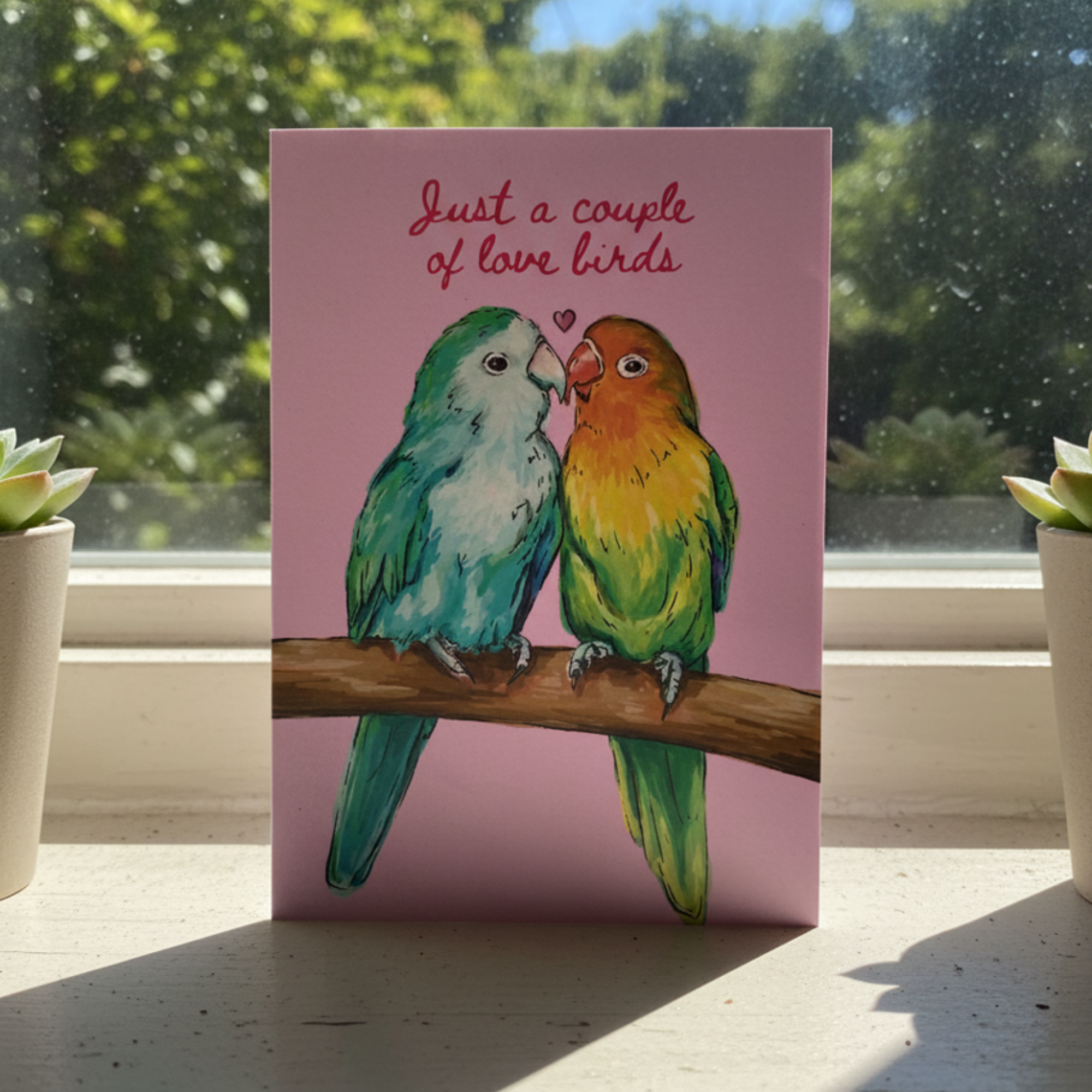 Card with two love birds on a branch and 'Just a couple of love birds' text, placed on a surface with a window and plants in the background.