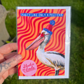 Card with a pelican wearing a party hat against a red wavy background, held by a person outdoors.