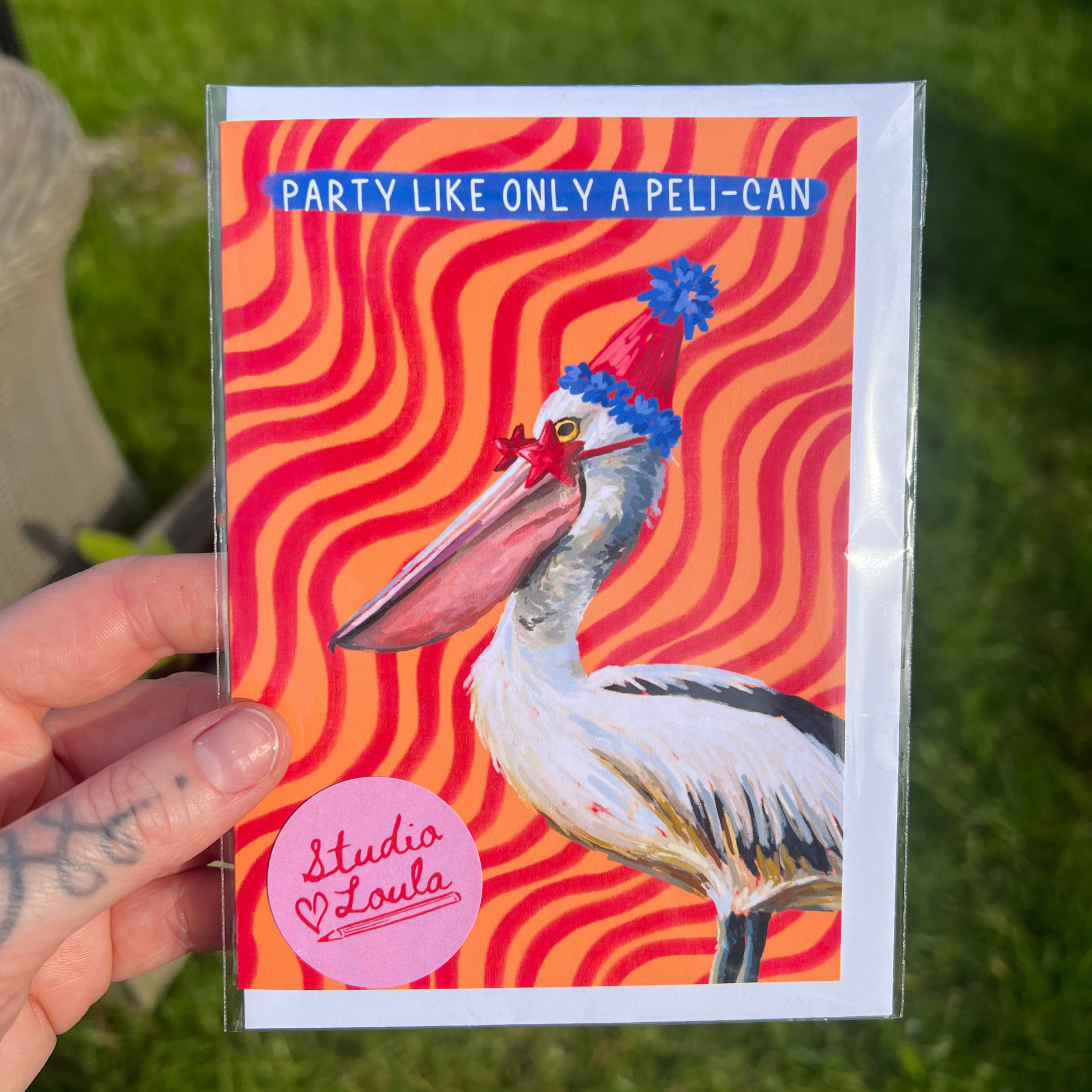 Card with a pelican wearing a party hat against a red wavy background, held by a person outdoors.