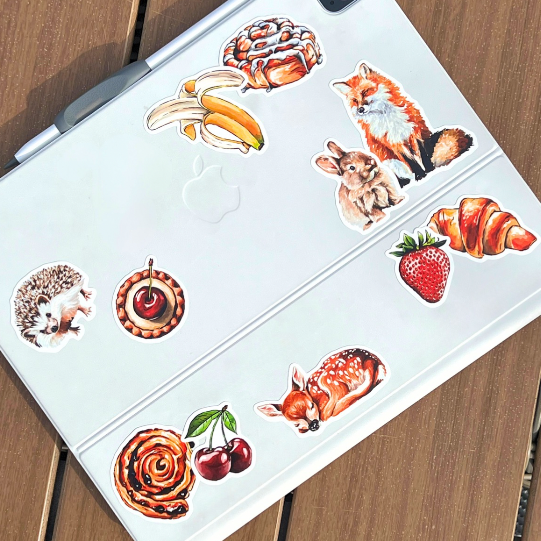 An Ipad cover decorated with colorful animal and food stickers, including a fox, rabbit, bananas, strawberries, croissants, cherries, and a hedgehog, placed on a wooden surface.