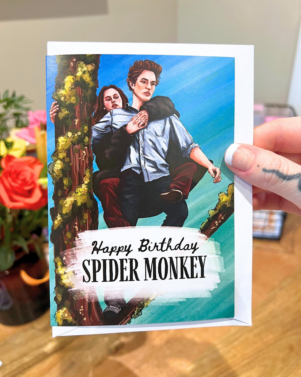 Birthday card with illustration of two people on a branch, text 'Happy Birthday SPIDER MONKEY', held by a hand.