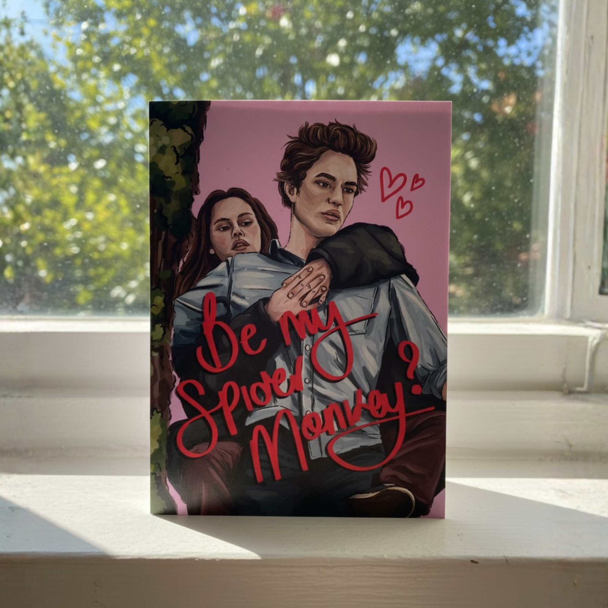 Card with illustration of two people and text 'Be my Spider Monkey?' on a pink background, placed on a windowsill.