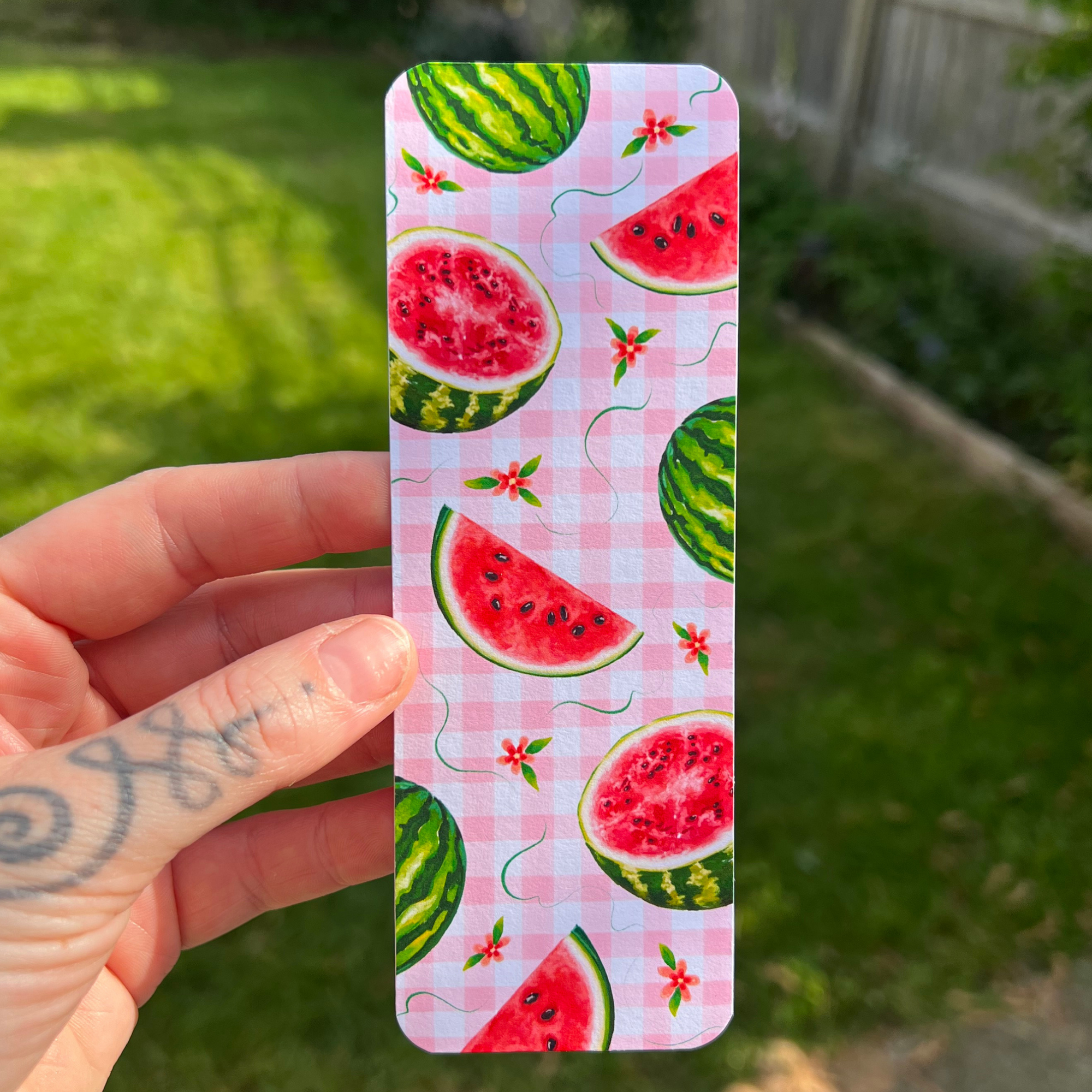 A hand with a tattoo holds a rectangular bookmark featuring a pink gingham background and a watermelon slice and whole watermelon pattern, against a blurred green backyard background.