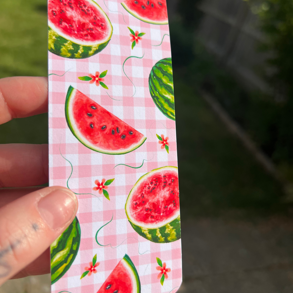 A hand holding a slim rectangular card or bookmark featuring a pink gingham pattern with watermelon slices, green rind, and small red flowers, against an outdoor blurred background.