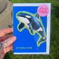 Card with an orca whale illustration and 'Studio Loula' branding held by a hand on grass.