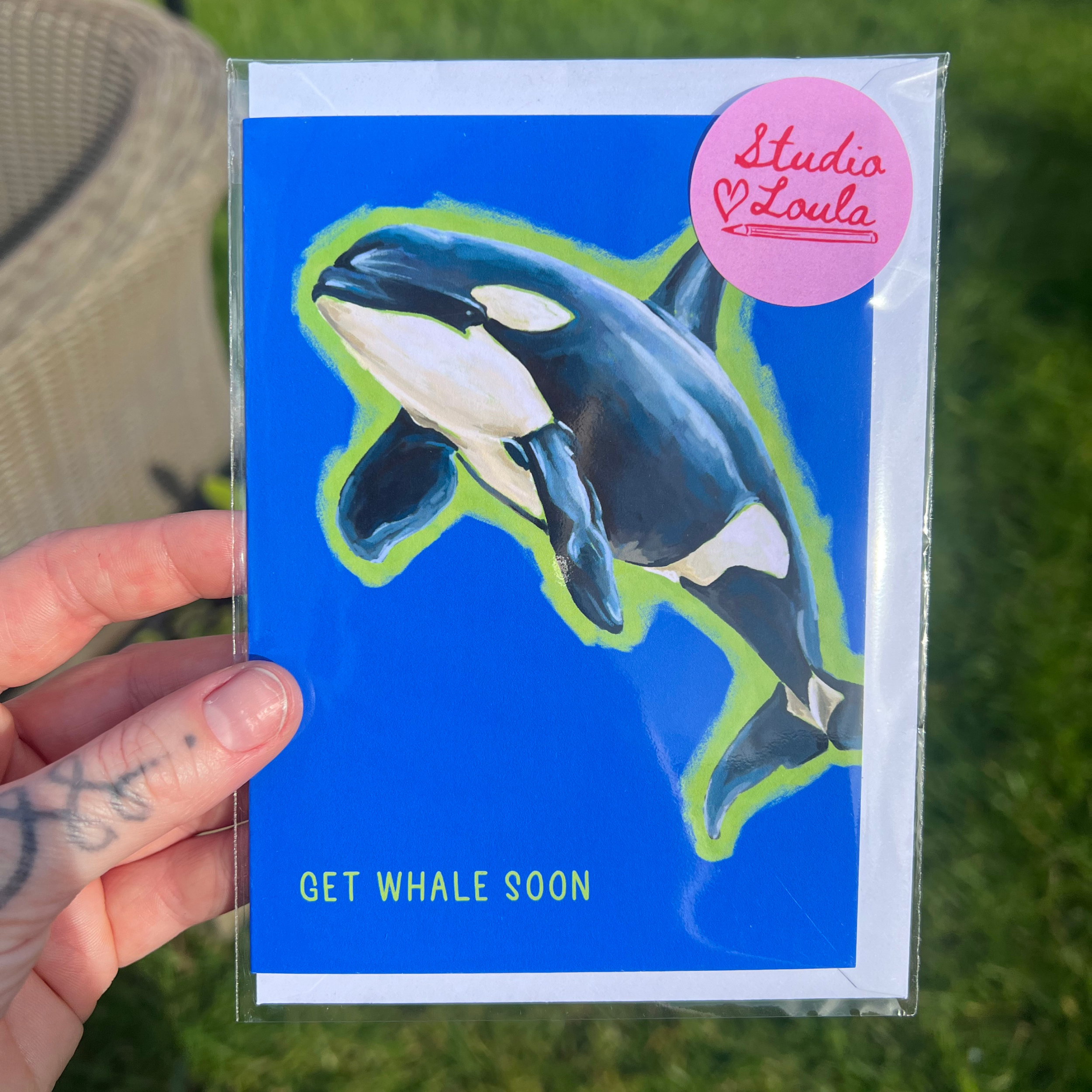 Card with an orca whale illustration and 'Studio Loula' branding held by a hand on grass.